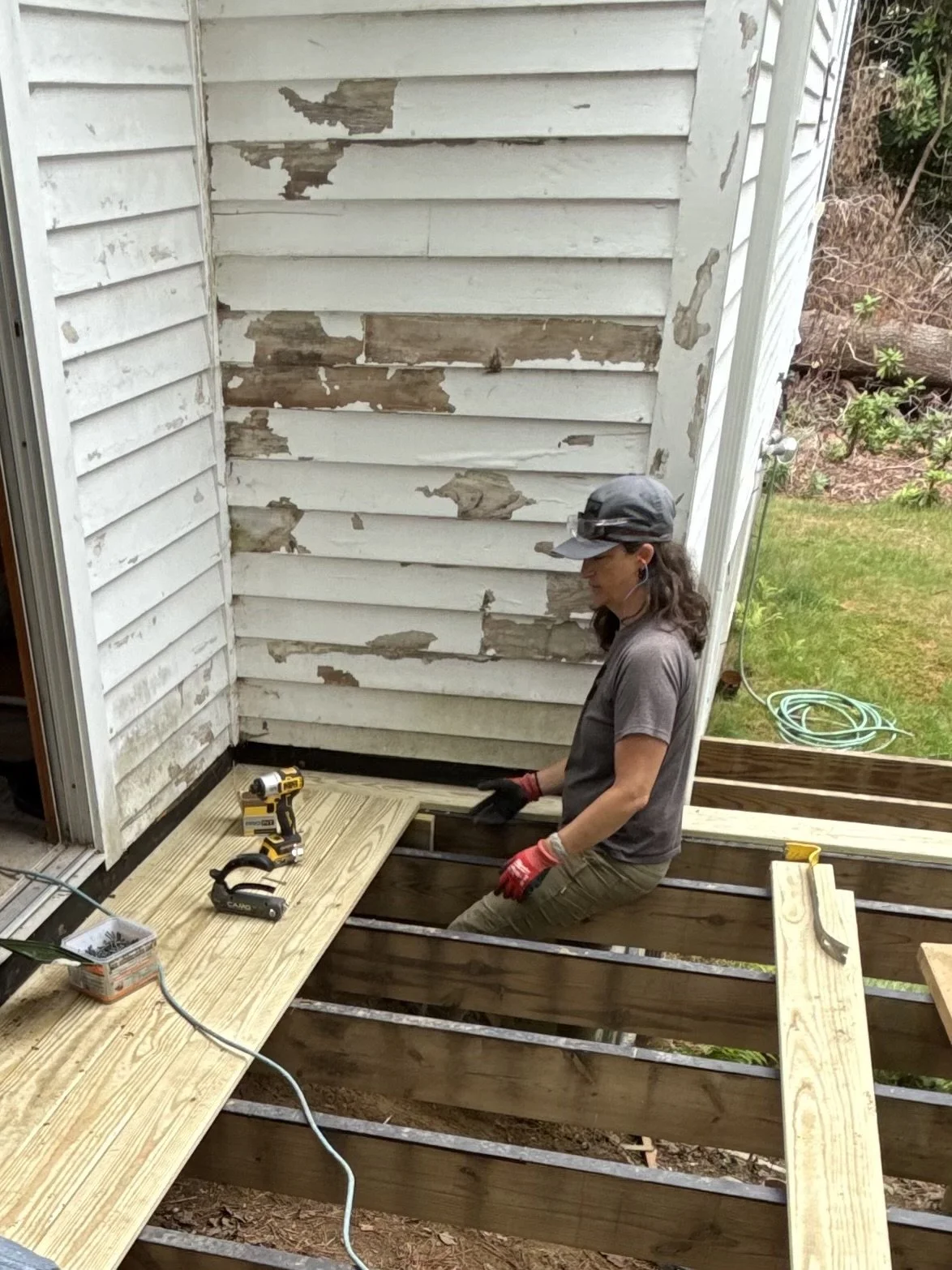 Woman working on a deck construction, sitting on the unfinished deck with drilled tools on the wooden surface, and a weathered, peeling white house exterior in the background.