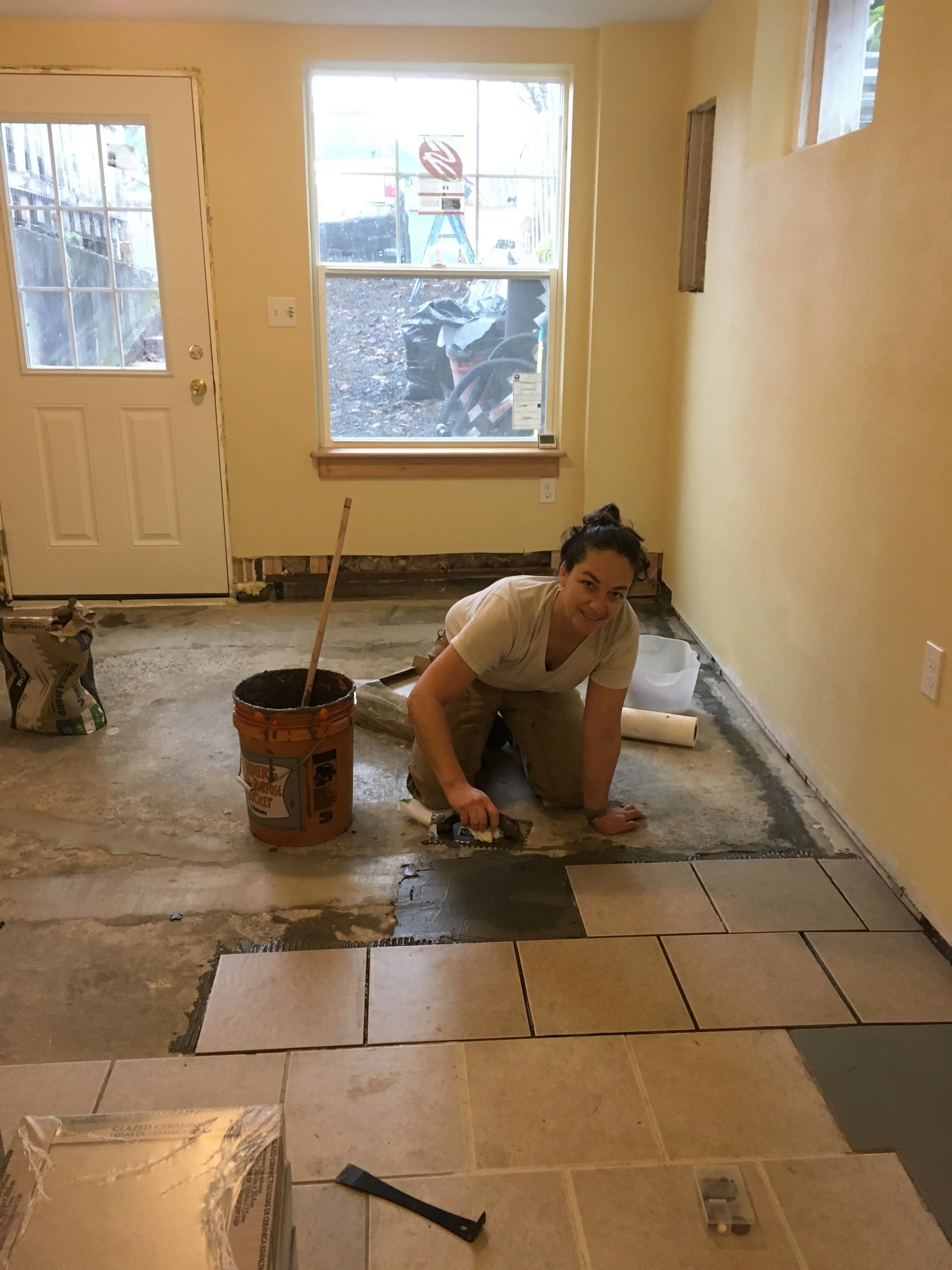 Woman installing tile flooring in a house during renovation, kneeling on the floor with a trowel, surrounded by construction materials and tools.
