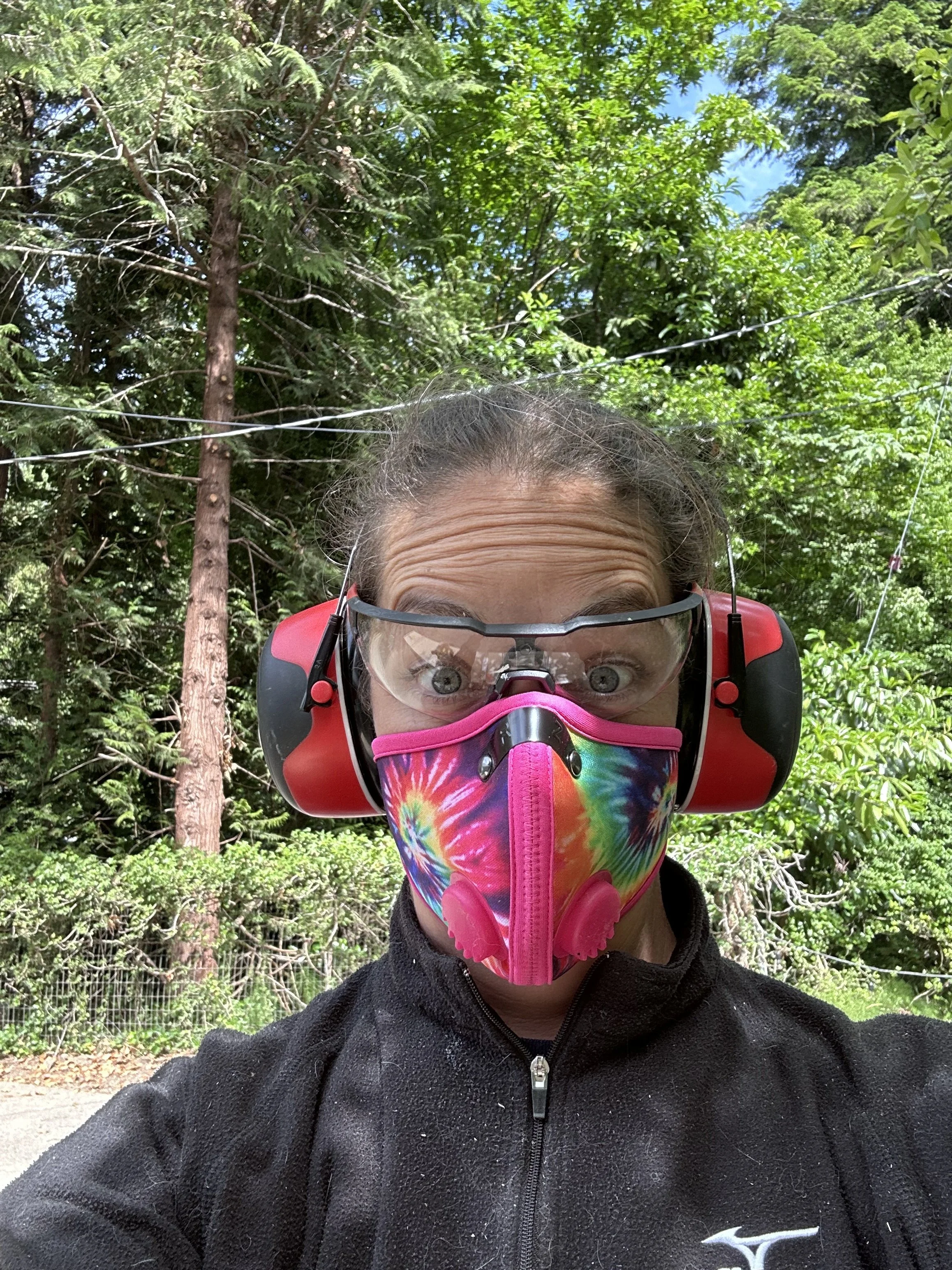 A woman wearing safety ear protection, a multicolored tie-dye face mask, and safety glasses takes a selfie outdoors in a wooded area with lush green trees and a blue sky overhead.