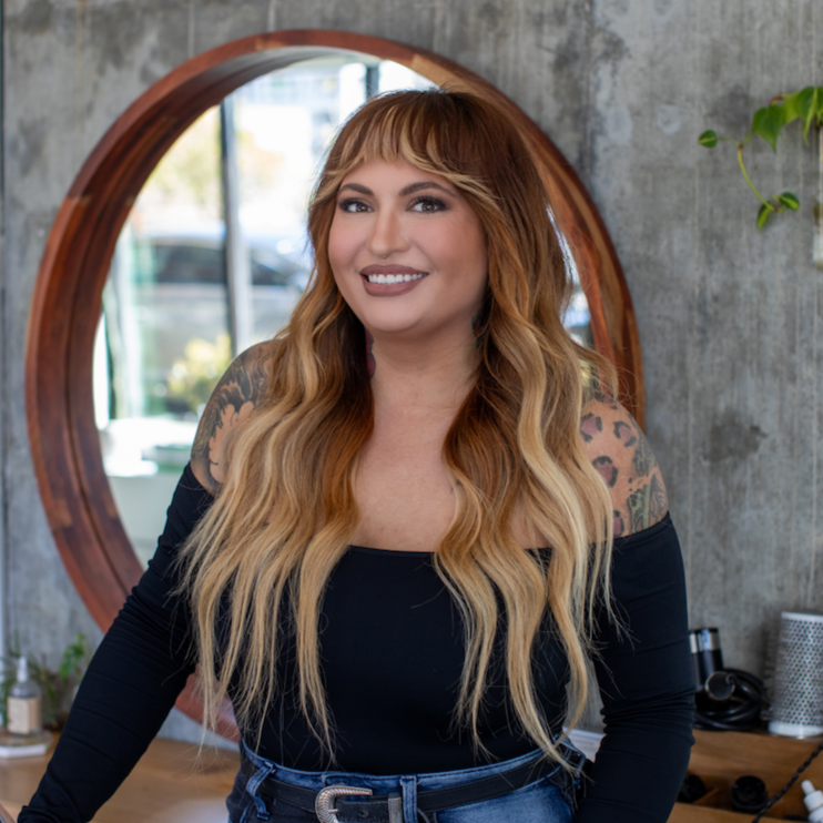 A smiling woman with long, wavy, blonde hair standing in a salon or studio. She has tattoos on her shoulders and wears a black off-the-shoulder top. Behind her is a round mirror on a concrete wall, and a plant is partially visible to the side.