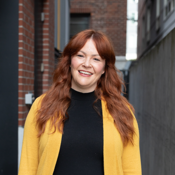 A woman with long red hair, smiling, wearing a black top and yellow cardigan, standing outdoors in an alley between brick and wooden buildings.