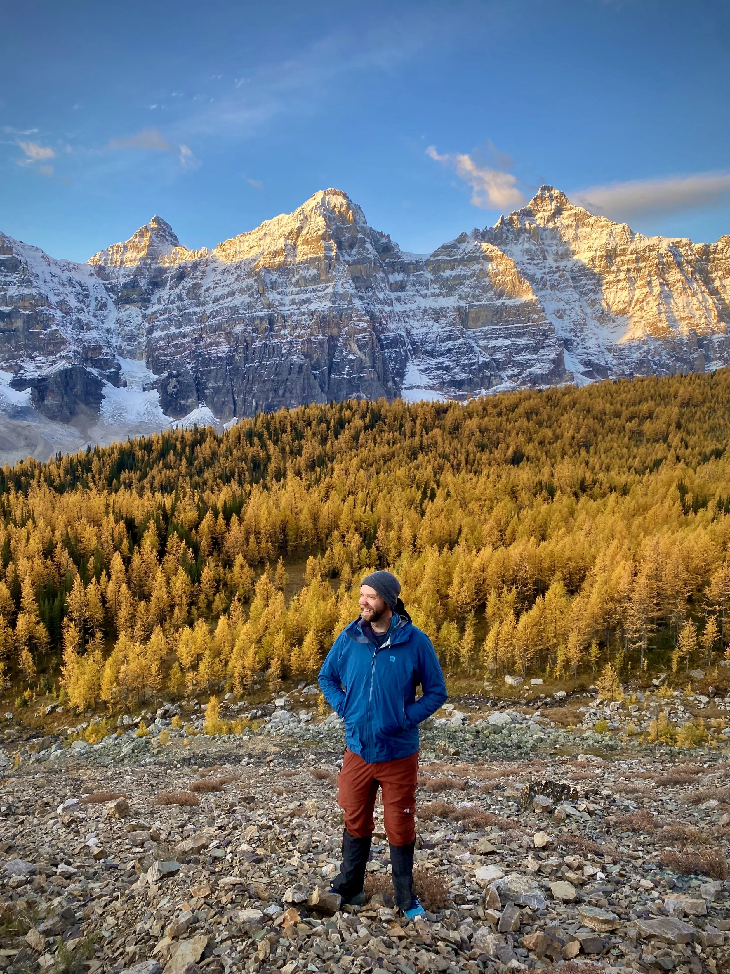 A man wearing a blue coat and red pants stands on a mountainside with larch trees and mountains behind him in Larch Valley in Banff National Park at sunrise.