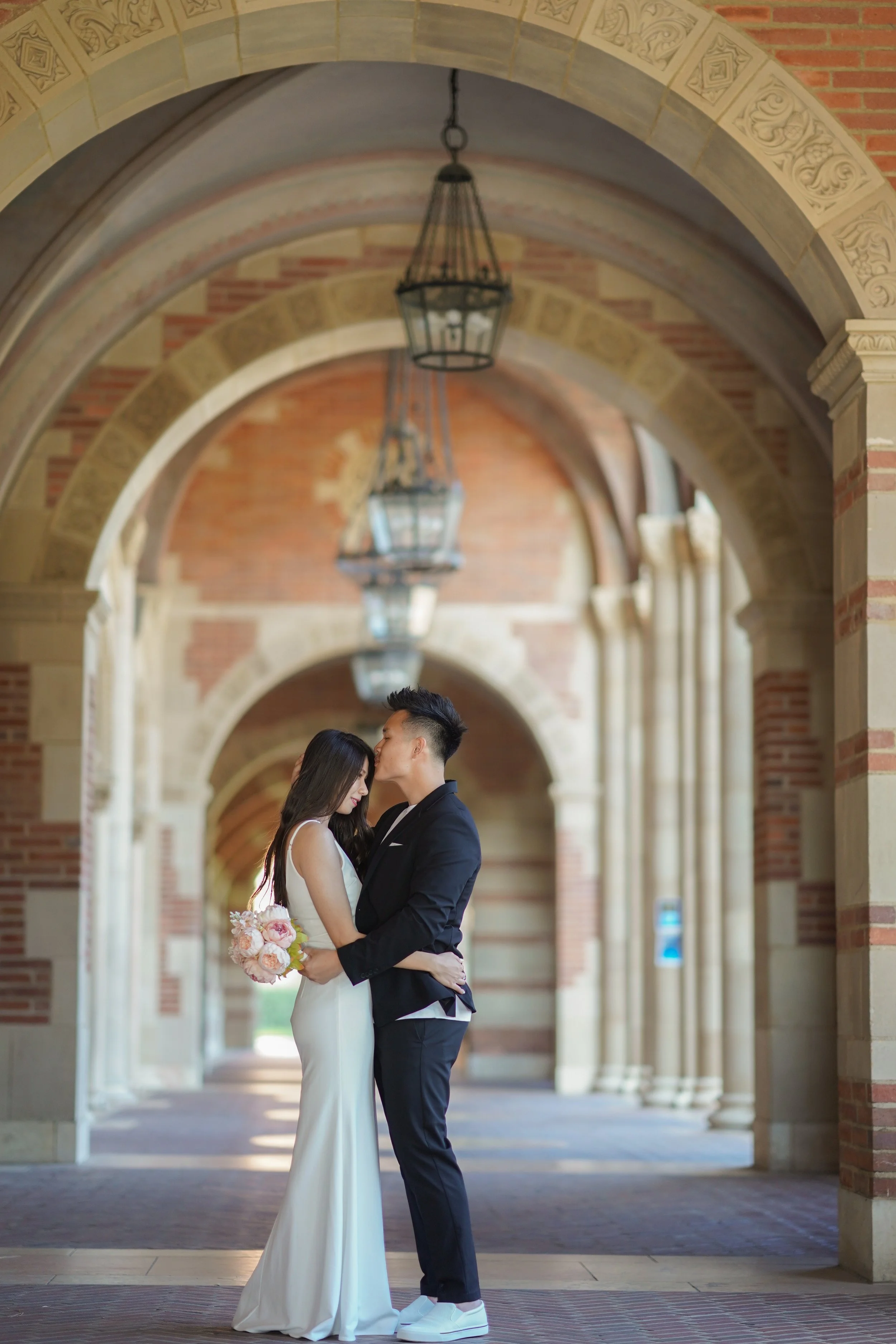 A bride and groom share a romantic kiss in an arched, brick and stone corridor with hanging lanterns.