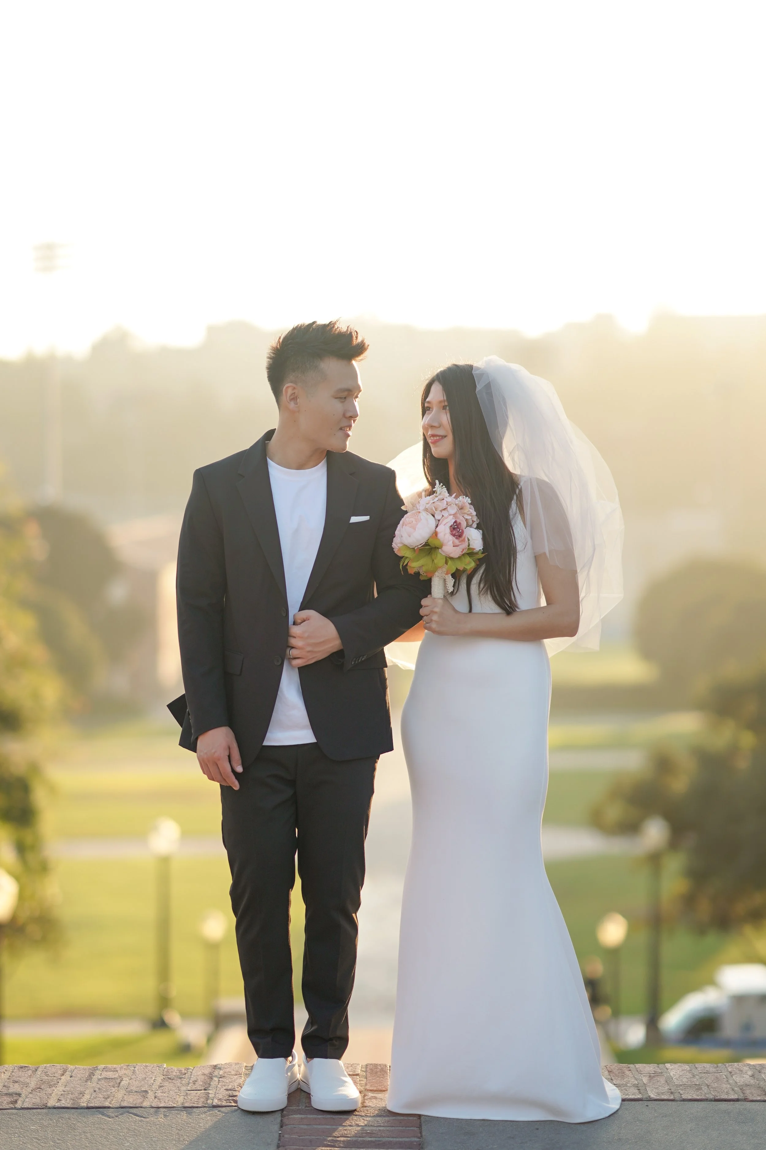 A couple dressed in wedding attire walking outdoors at sunset, with the woman holding a bouquet of pink and white flowers.