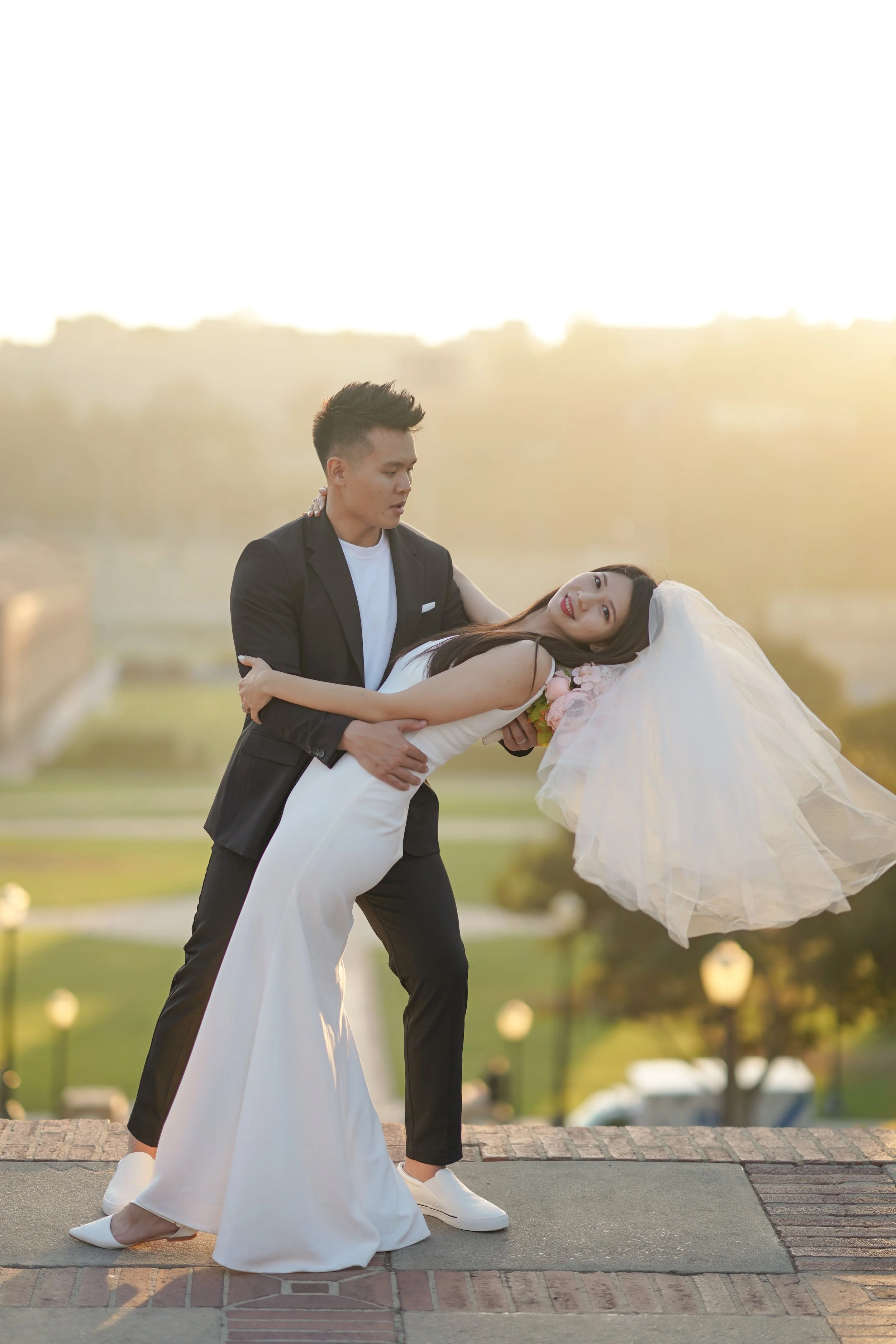 A couple in wedding attire dancing outdoors at sunset, with the man in a black suit and the woman in a white wedding gown and veil, on a brick surface with a park and trees in the background.
