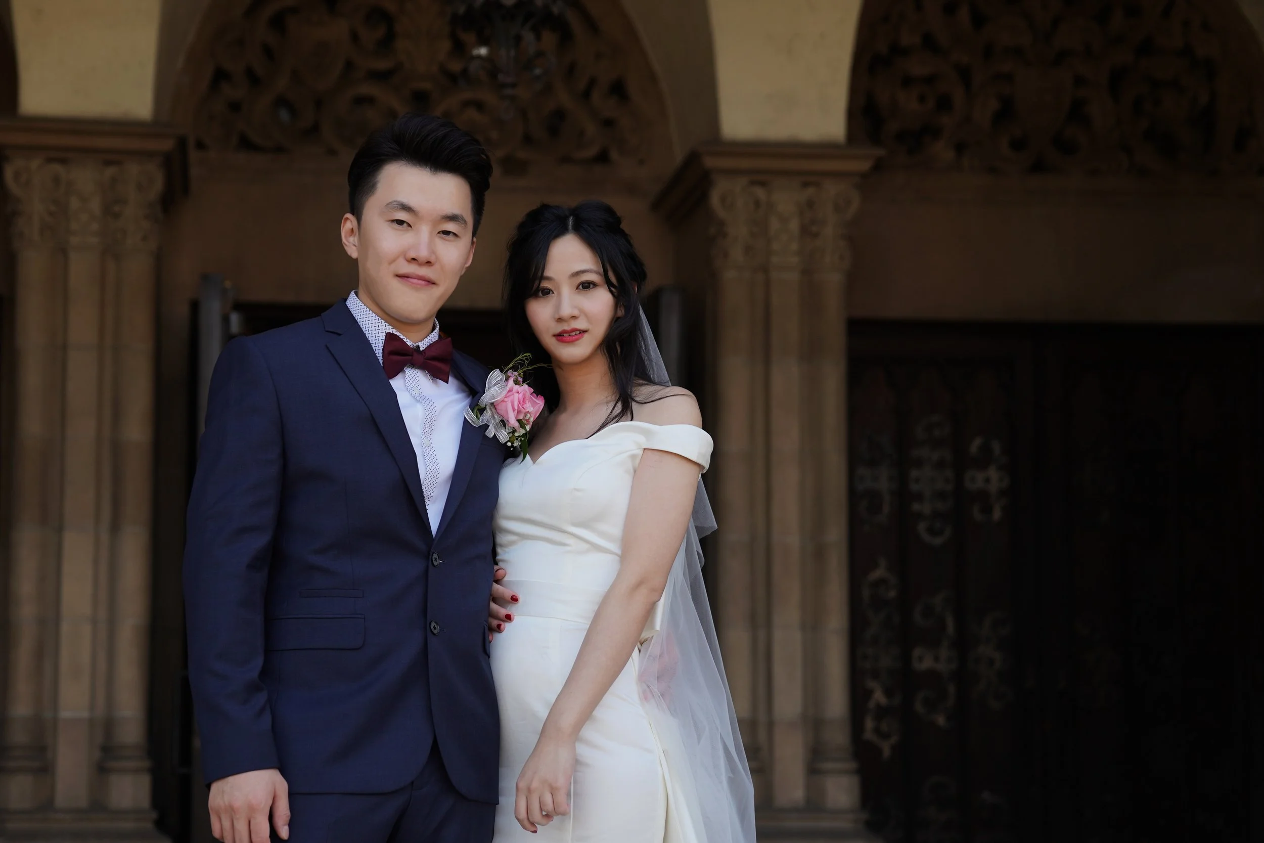 A newlywed couple dressed in wedding attire standing in front of an ornate building, the groom in a navy suit with a bow tie and boutonniere, and the bride in a white dress with a veil.