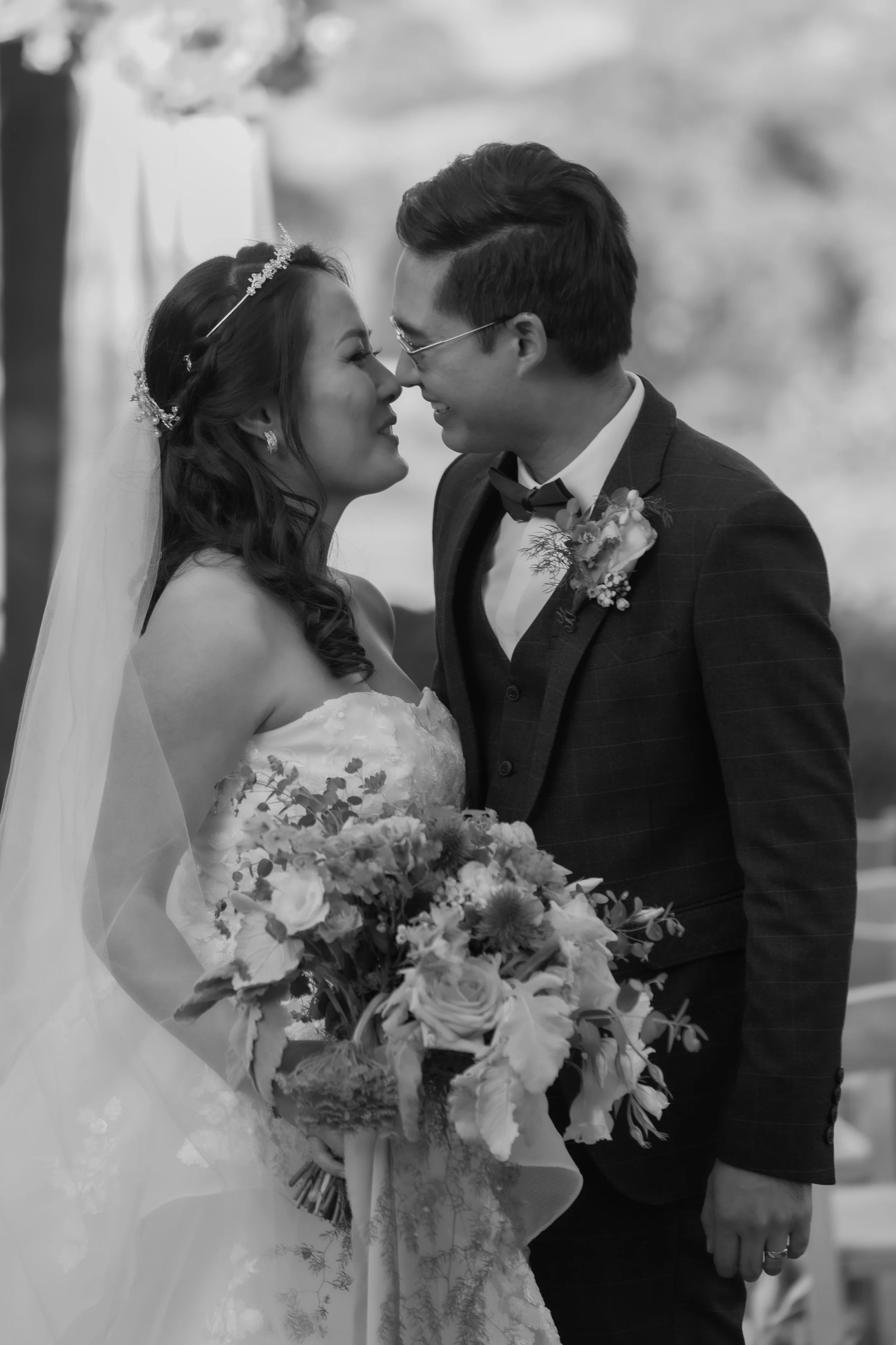 Black and white photo of a bride and groom with their foreheads touching, smiling, during their wedding ceremony. The bride is wearing a strapless lace gown, a veil, and holding a large bouquet of flowers. The groom is dressed in a dark suit with a b
