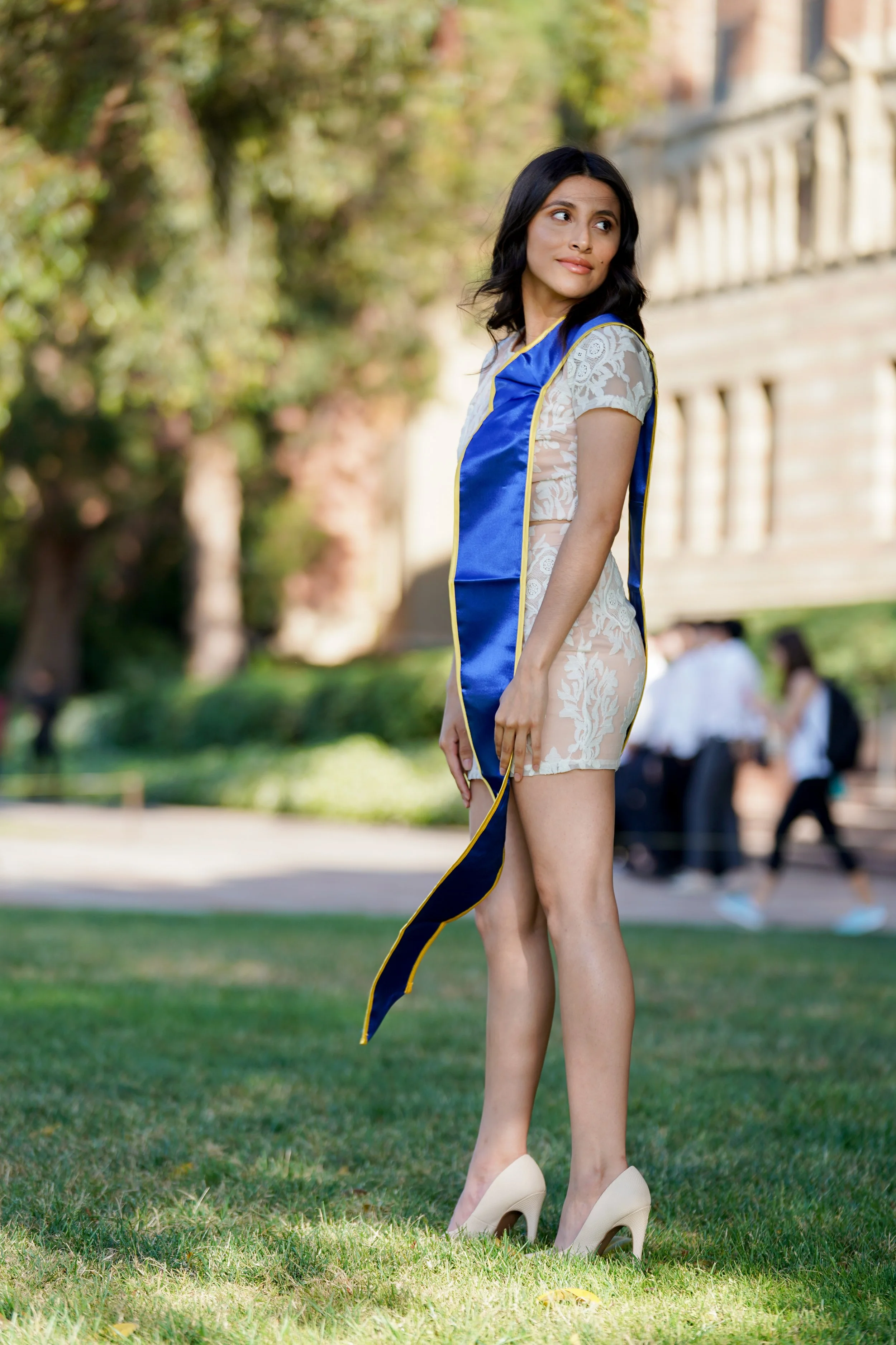 A woman wearing a blue graduation stole over a white lace dress standing on a grassy area outdoors during daytime. She is wearing high heels and appears to be at a graduation ceremony or celebration.