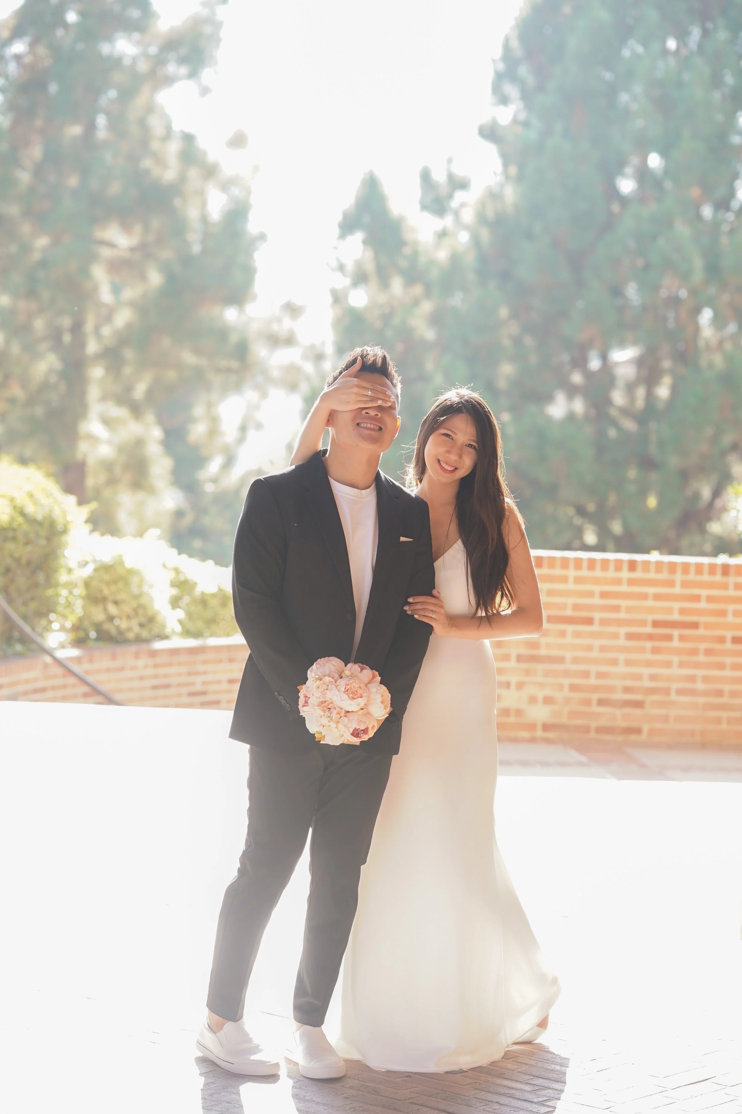 A couple at a wedding, with the groom holding a bouquet of pink flowers and the bride smiling. The groom covers his eyes with one hand, while the bride has her arm around him. They are outdoors with trees in the background.