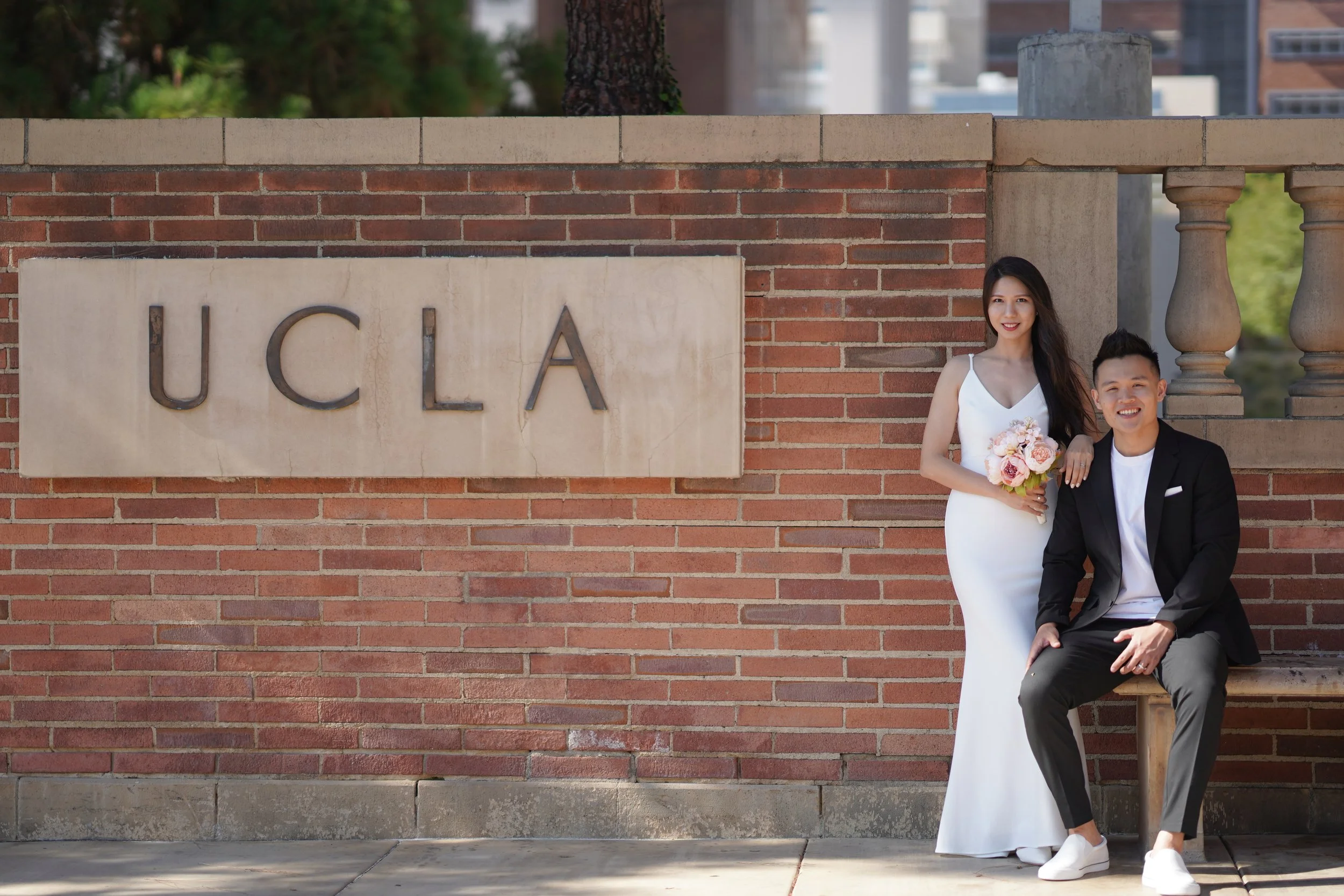A woman in a white dress standing with a man in a black suit, sitting on a bench, in front of a brick wall with a sign that says UCLA.