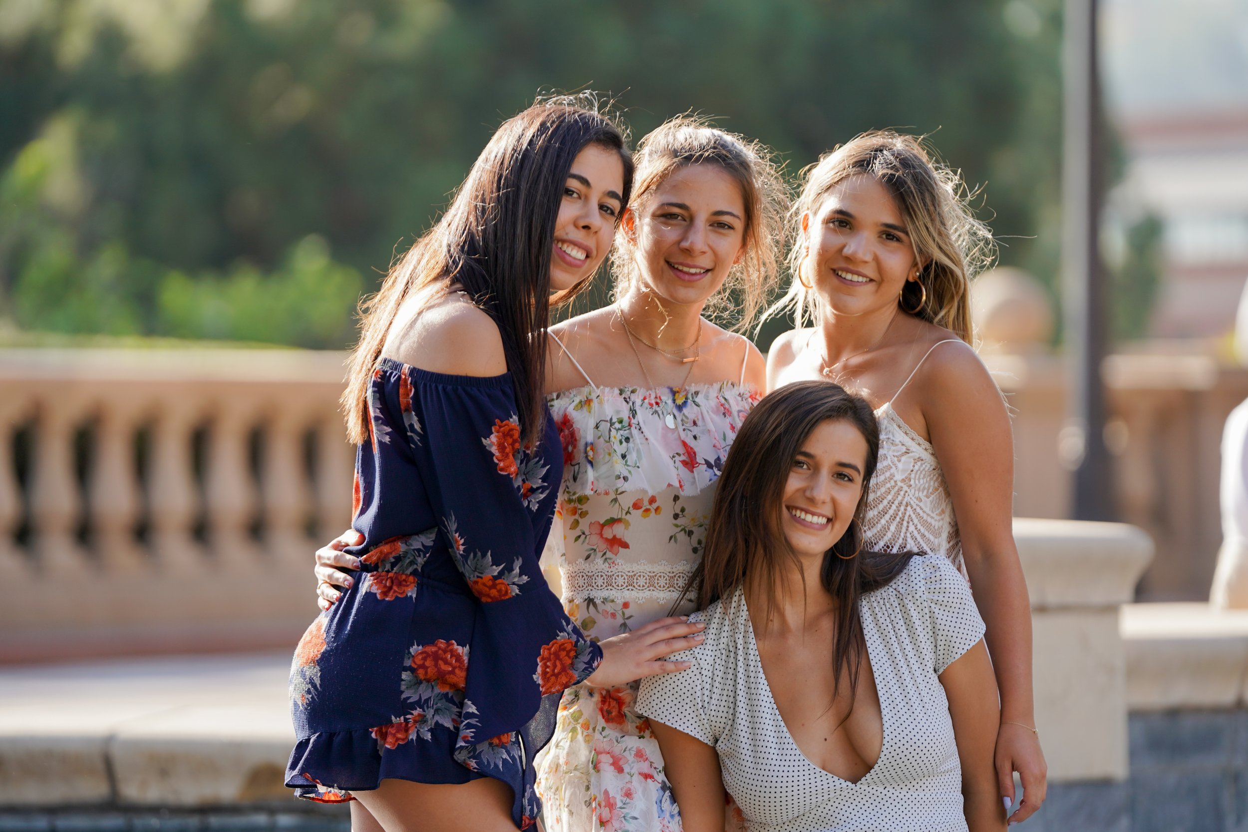 Five young women posing together outdoors, smiling at the camera, with a blurred background of trees and a stone balustrade.