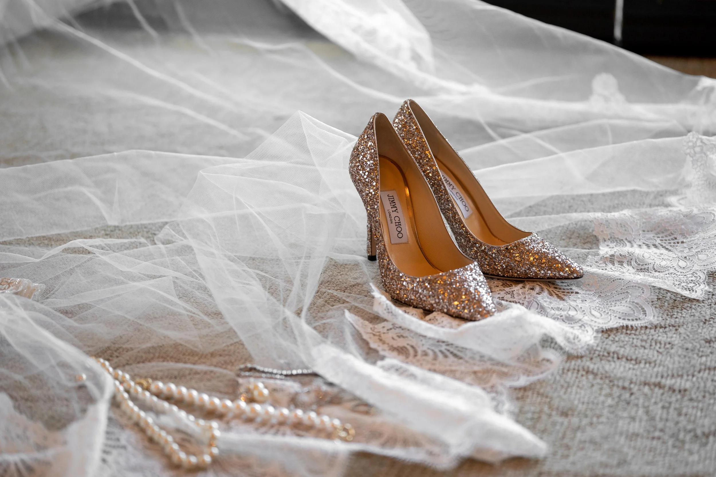 A pair of glittery Jimmy Choo high-heeled shoes surrounded by lace, tulle, and pearl necklaces on a textured surface.