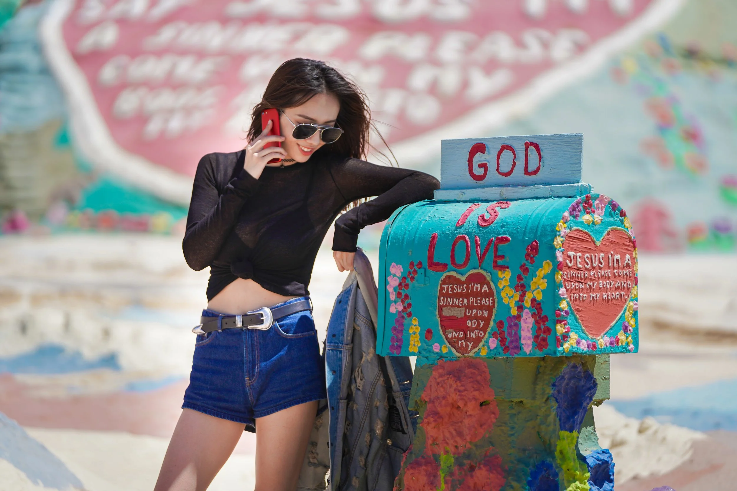 A woman in sunglasses and a black long-sleeve shirt talking on a red phone at Salvation Mountain with colorful painted signs and the message 'God is Love' on a blue mailbox-shaped structure.