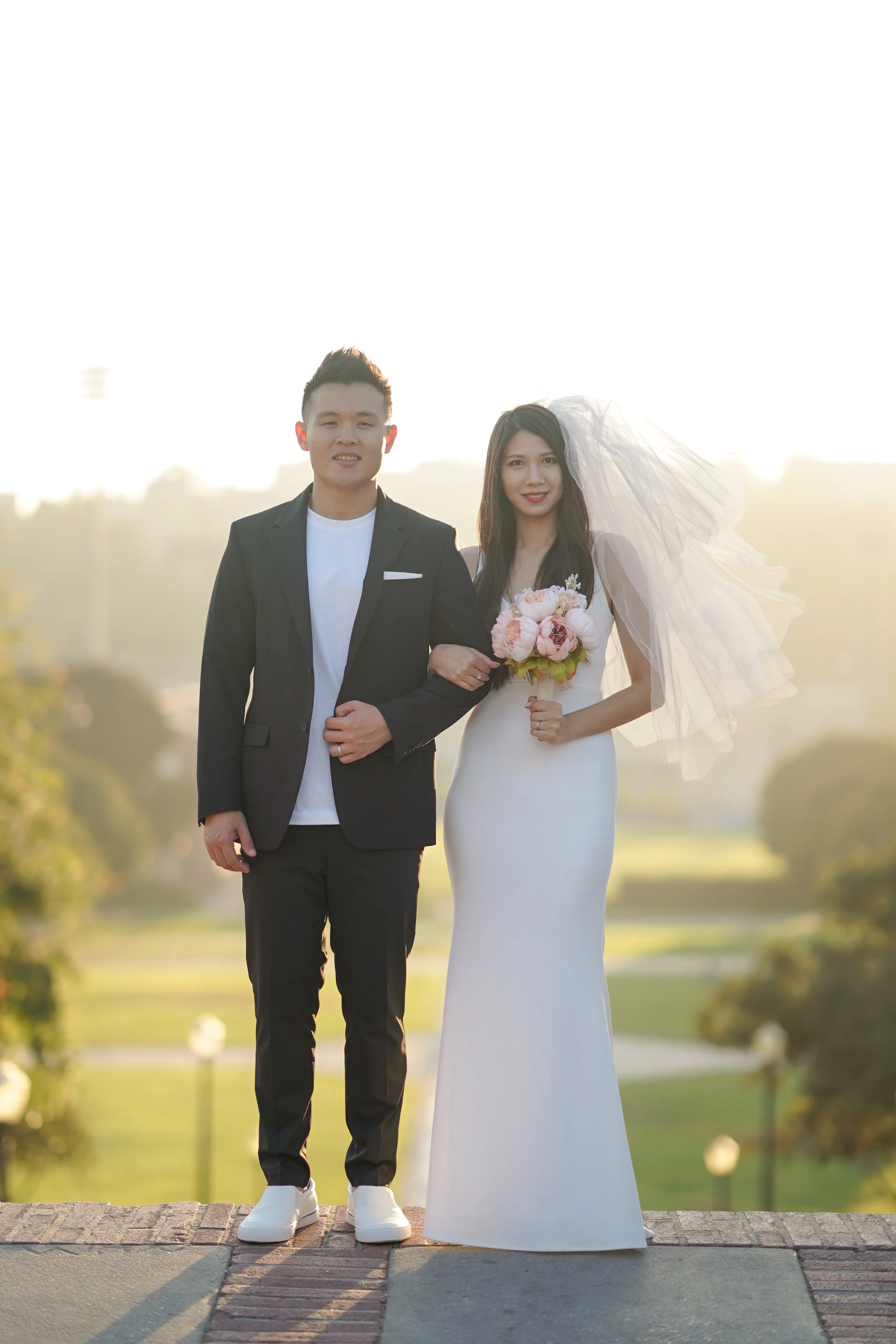 A bride and groom standing together outdoors during sunset, with the groom in a black suit and white shoes, and the bride in a white wedding gown holding a bouquet of pink and white flowers, the bride wearing a veil.