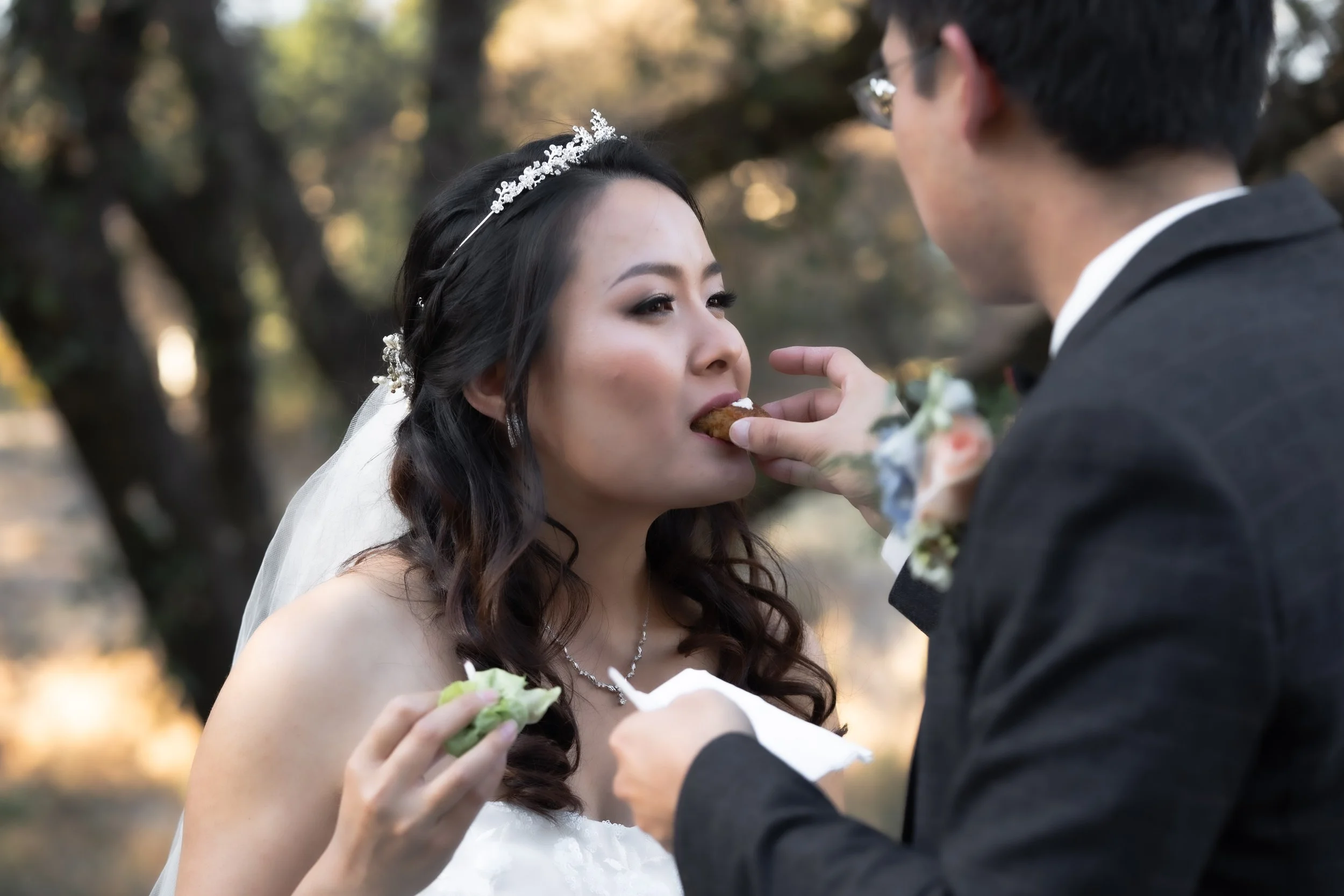 A bride and groom sharing a moment outdoors during their wedding, with the groom feeding the bride a piece of food.