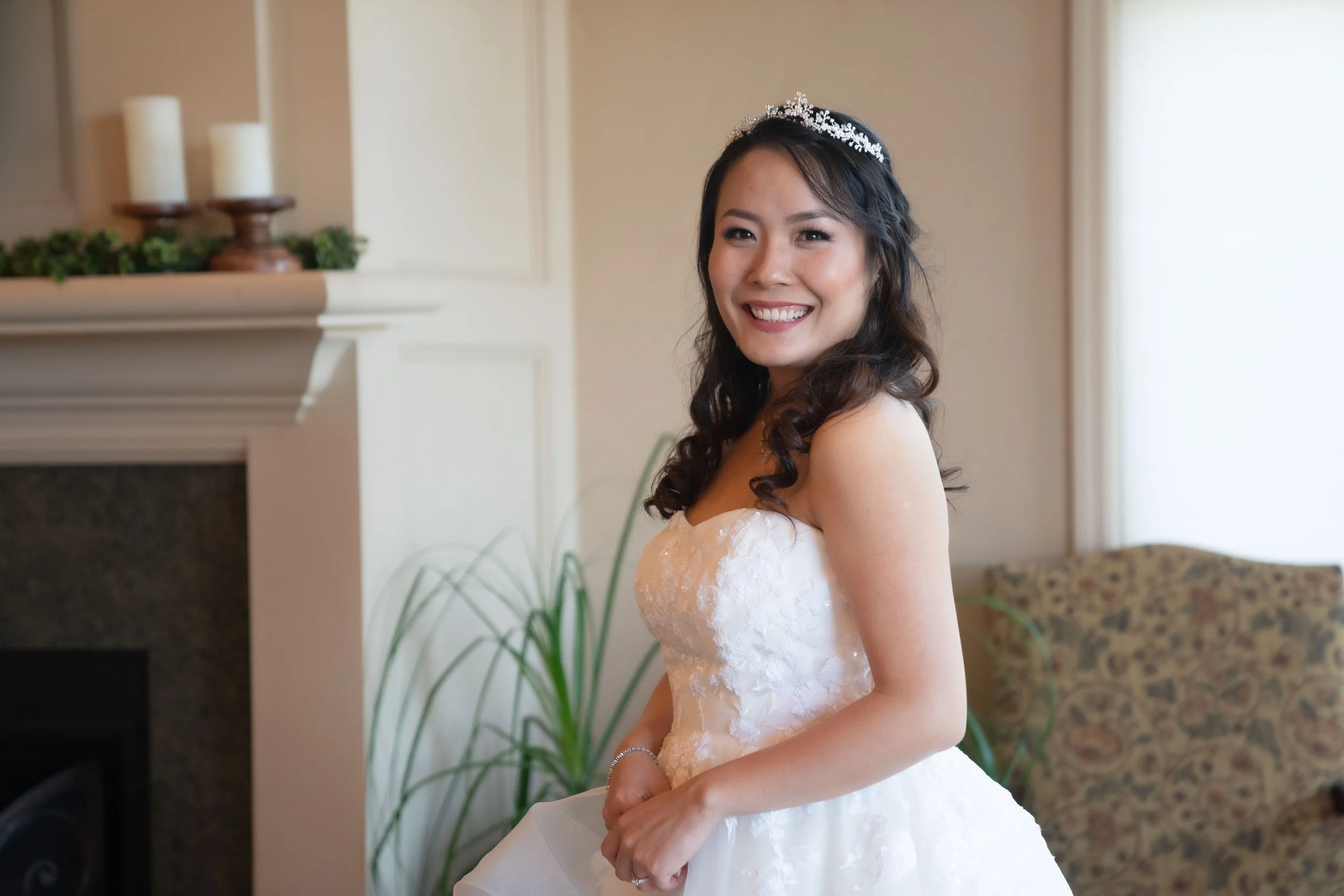 A young woman smiling in a white wedding gown with a tiara, standing indoors near a fireplace and a window.