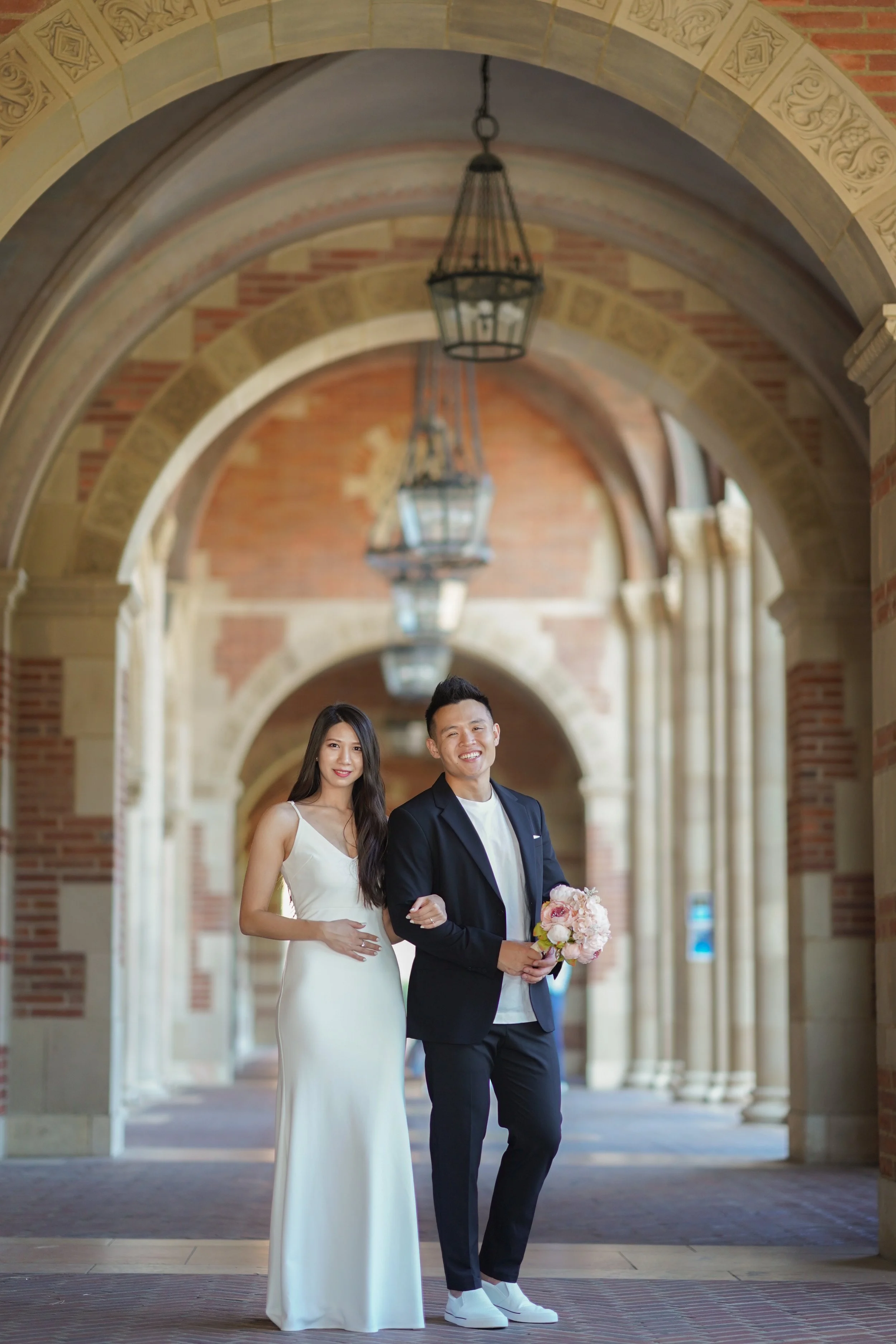 A happy couple, the woman in a white wedding dress and the man in a black suit with sneakers, standing under ornate brick and stone archways with hanging lanterns, holding a bouquet of pink and white flowers.