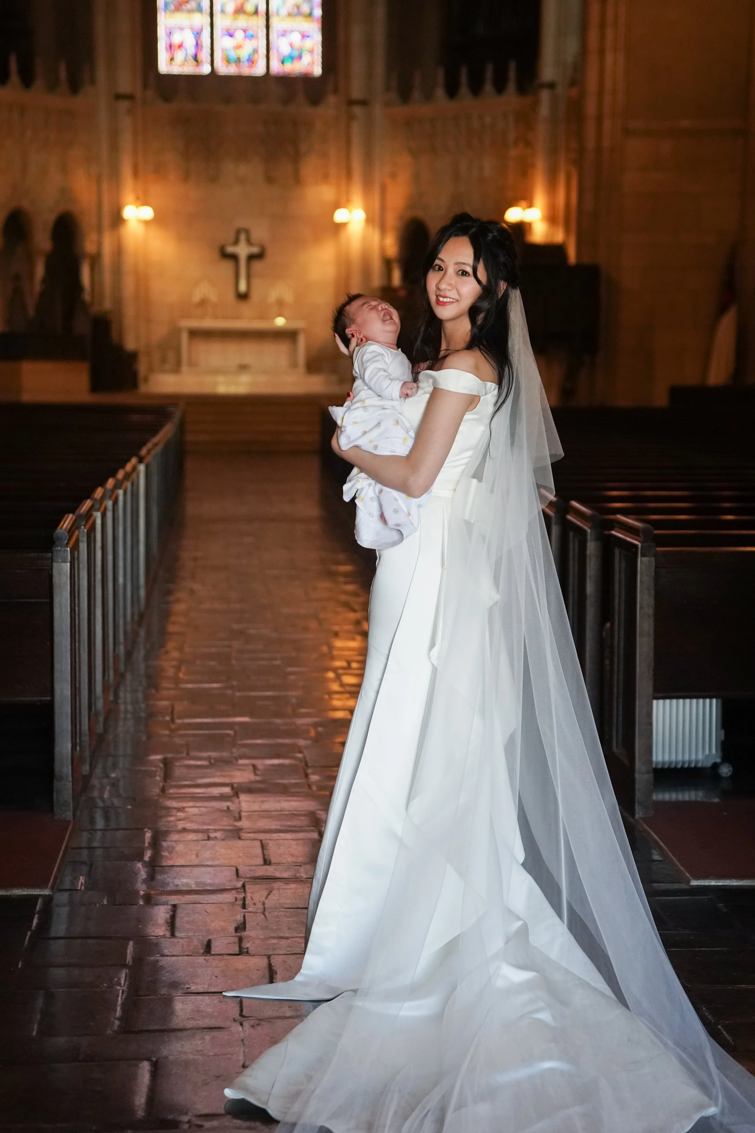 A woman in a white wedding dress holding a crying baby inside a church with wooden pews and stained glass windows.