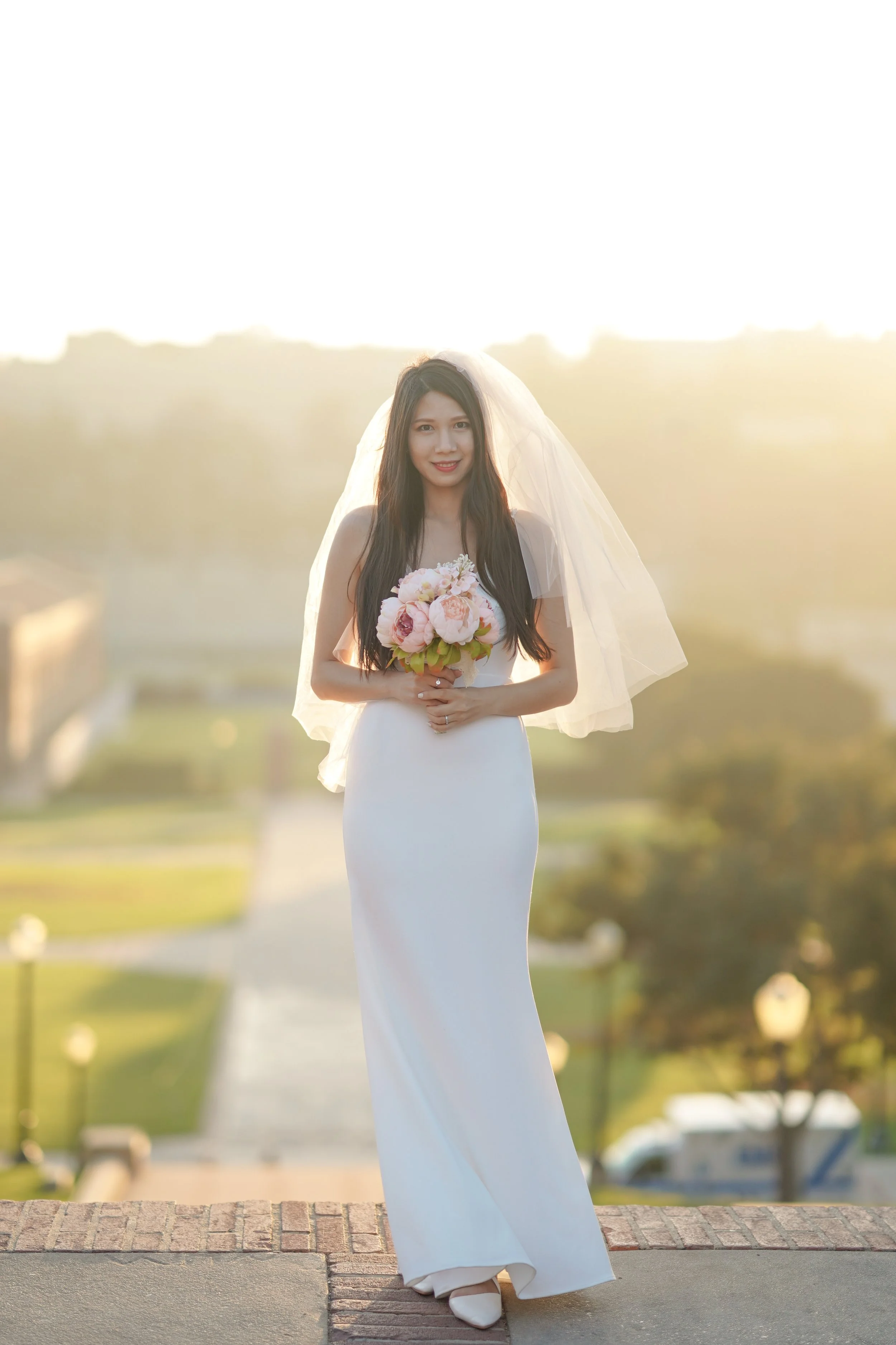 A bride in a white wedding gown holding a bouquet of pink and white flowers, standing outdoors at sunset with a blurred background of a park or garden.