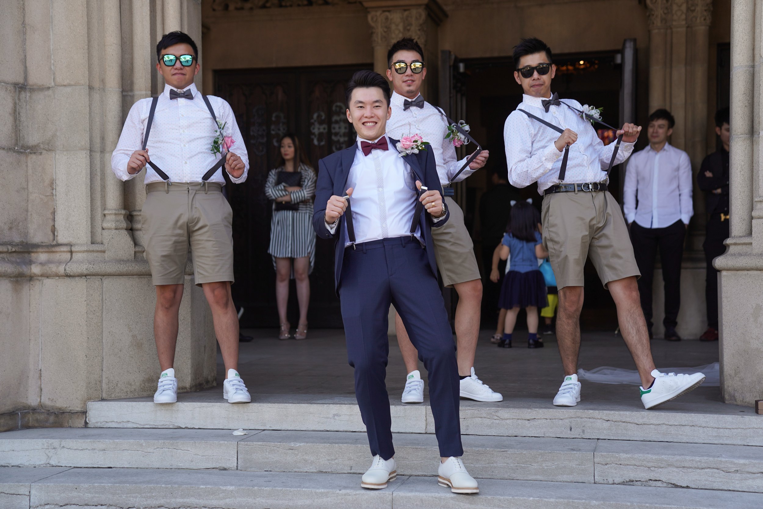A group of men posing on the steps of a church, dressed in semi-formal attire, with the man in the front wearing a navy blue suit and bow tie. The men in the background are wearing light shirts, ties, suspenders, and khaki shorts, with some wearing s