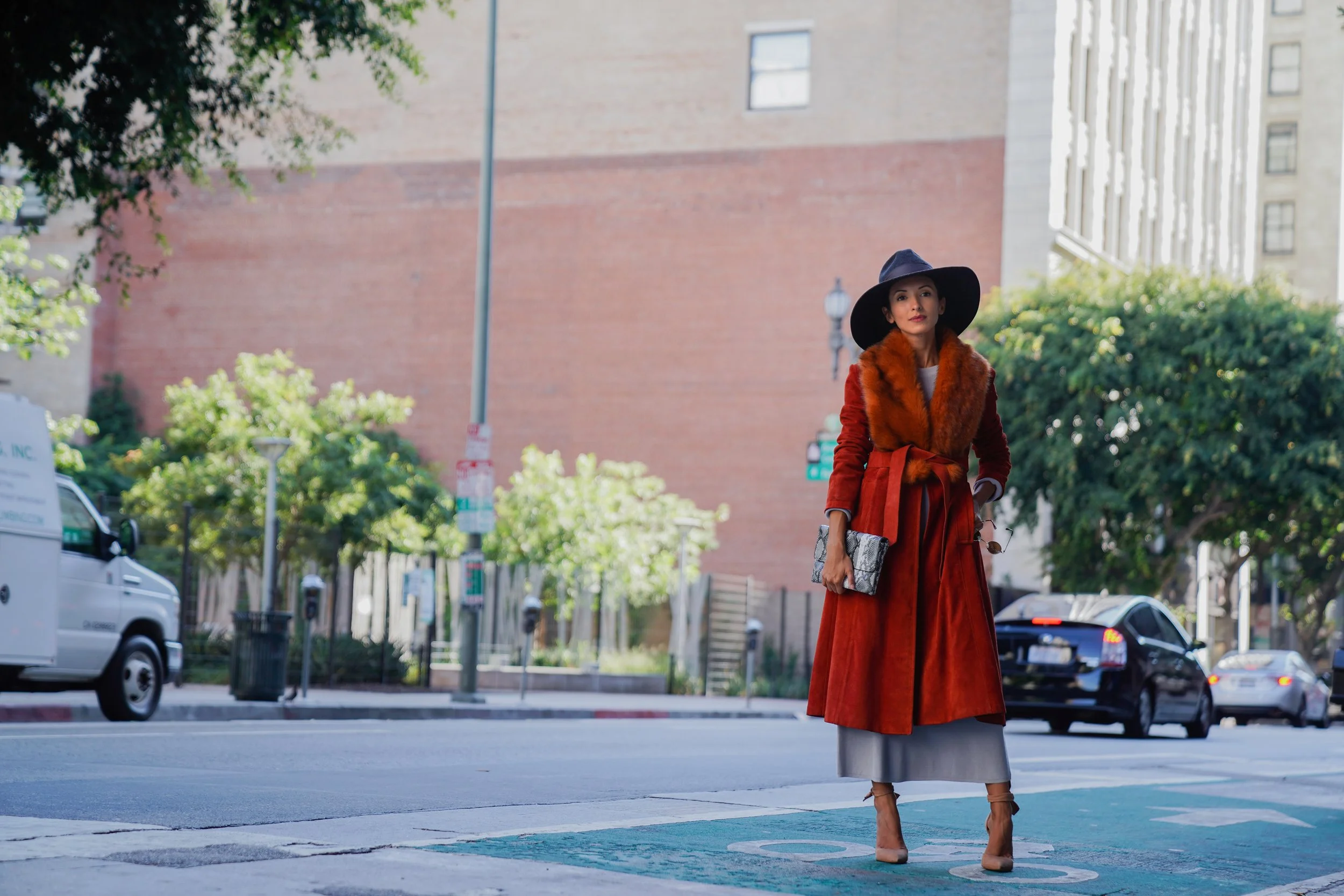 A woman in a red coat and gray skirt walking on a city sidewalk, wearing a large black hat and carrying a patterned clutch purse.