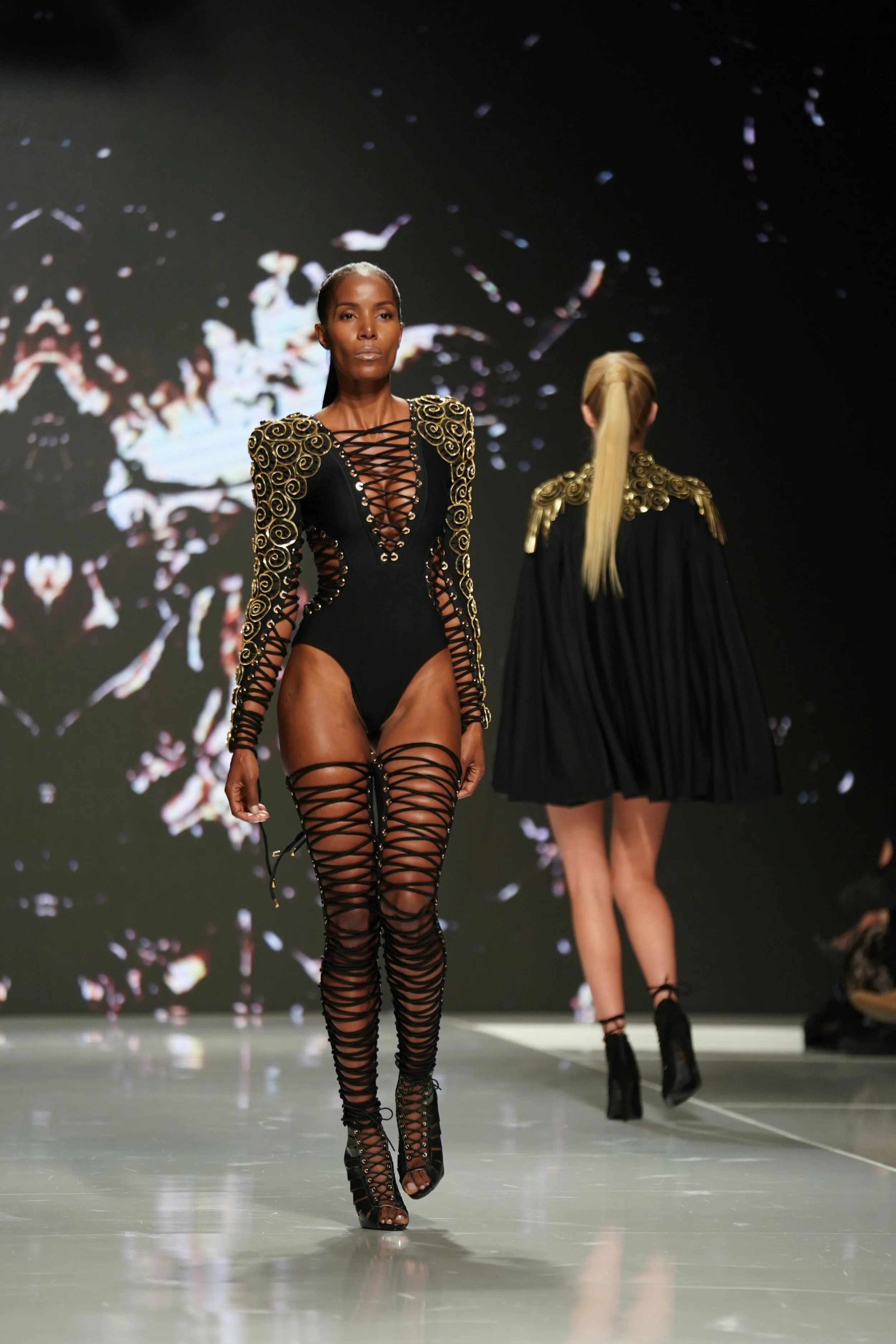 Model walking down runway in black and gold ornate fashion with lace-up details.