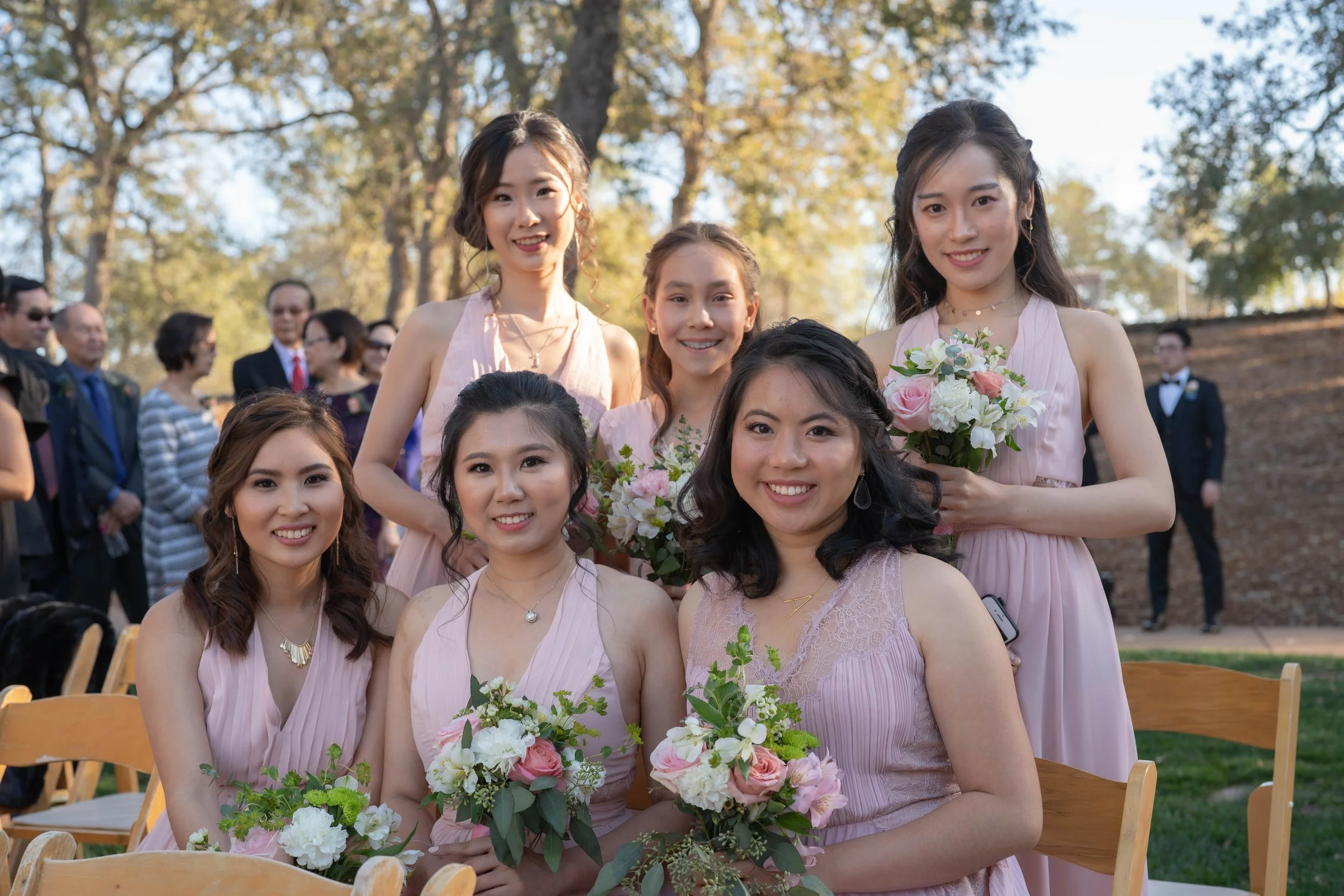 Six women in pink dresses holding bouquets of flowers, gathered outdoors during a sunny late afternoon, smiling for the camera at a social event or celebration.
