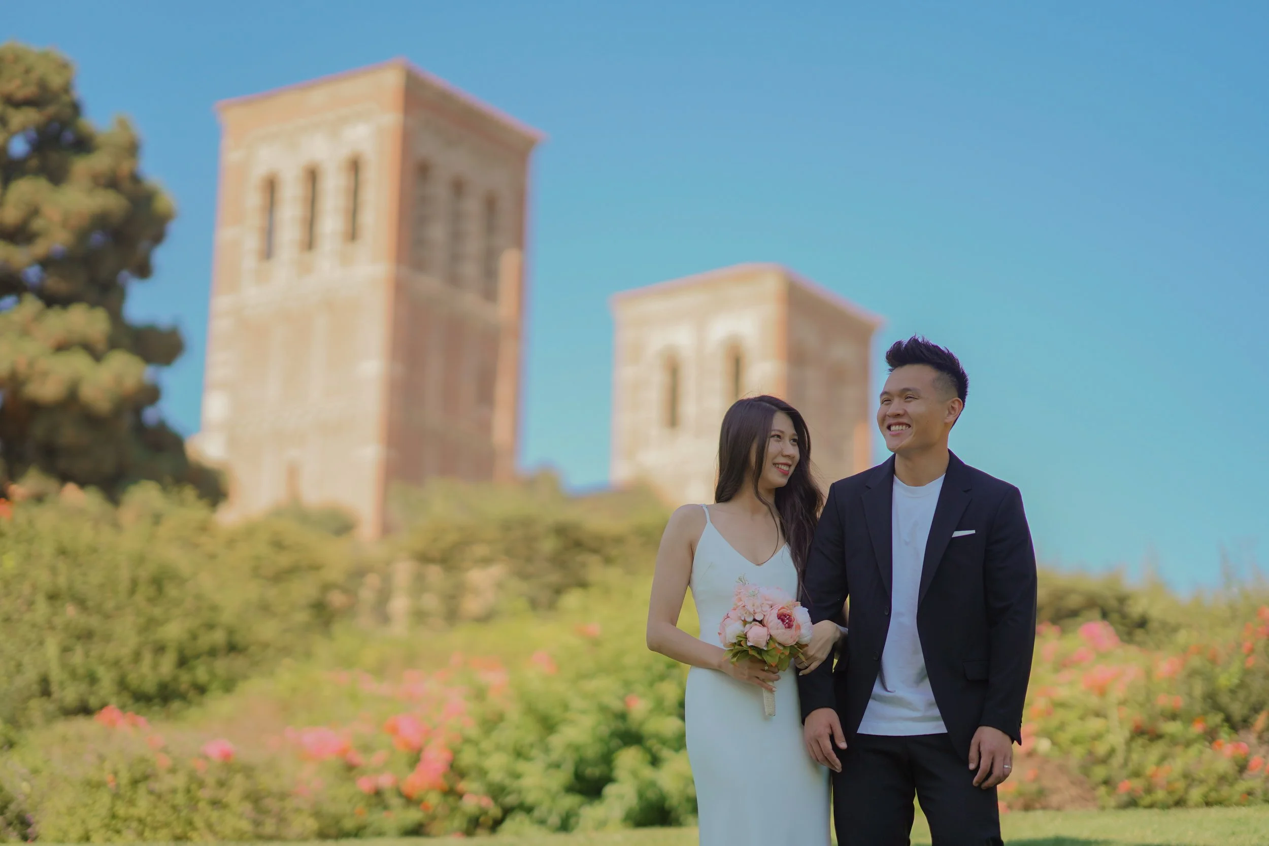 A couple dressed in wedding attire walking outdoors, holding hands, with a woman carrying a bouquet, smiling, and in the background there are two tall, historic brick towers under a clear blue sky, surrounded by green bushes and pink flowers.