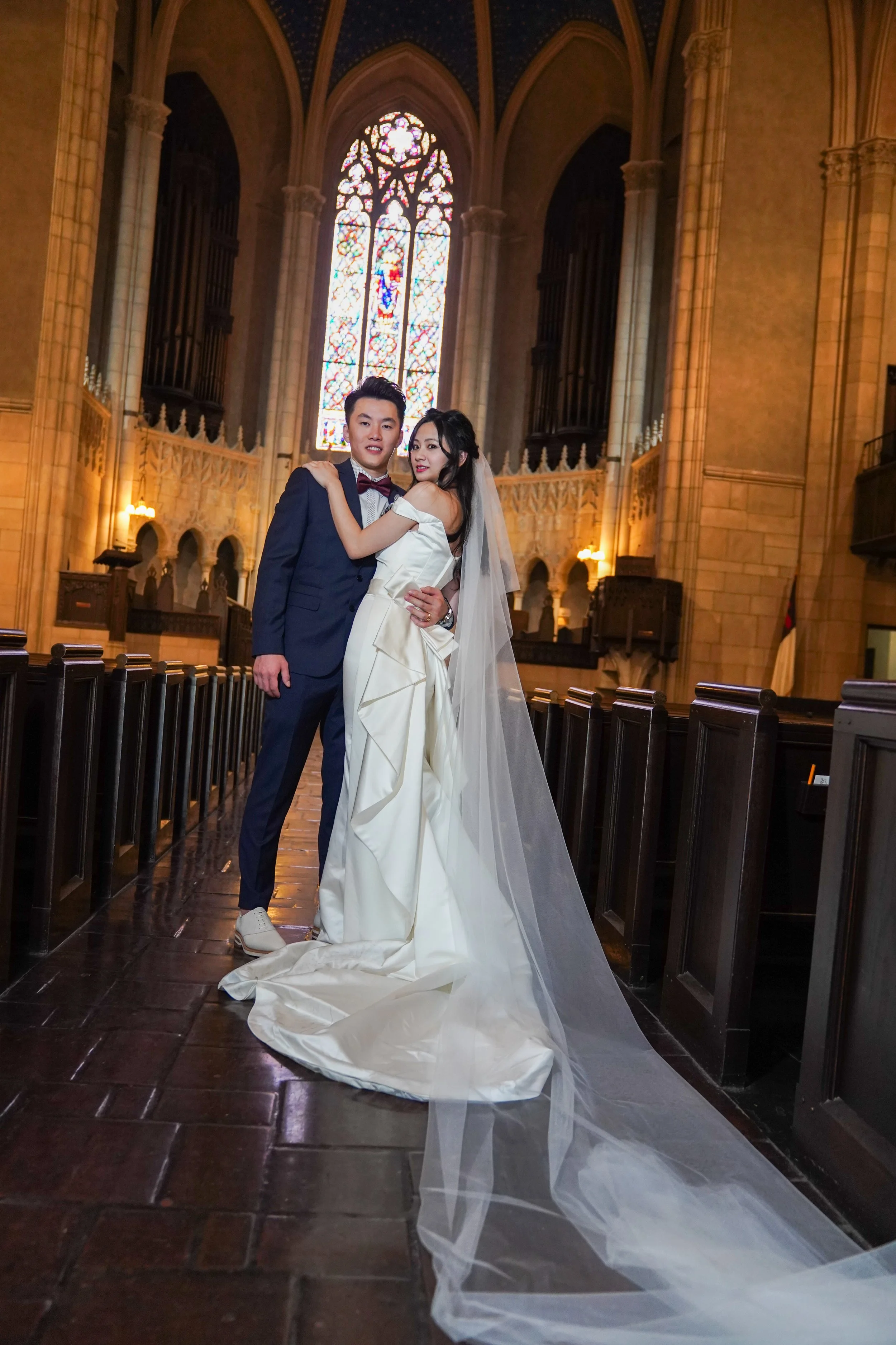 A bride and groom pose together inside a church with stained glass windows, wooden pews, and gothic architectural details.