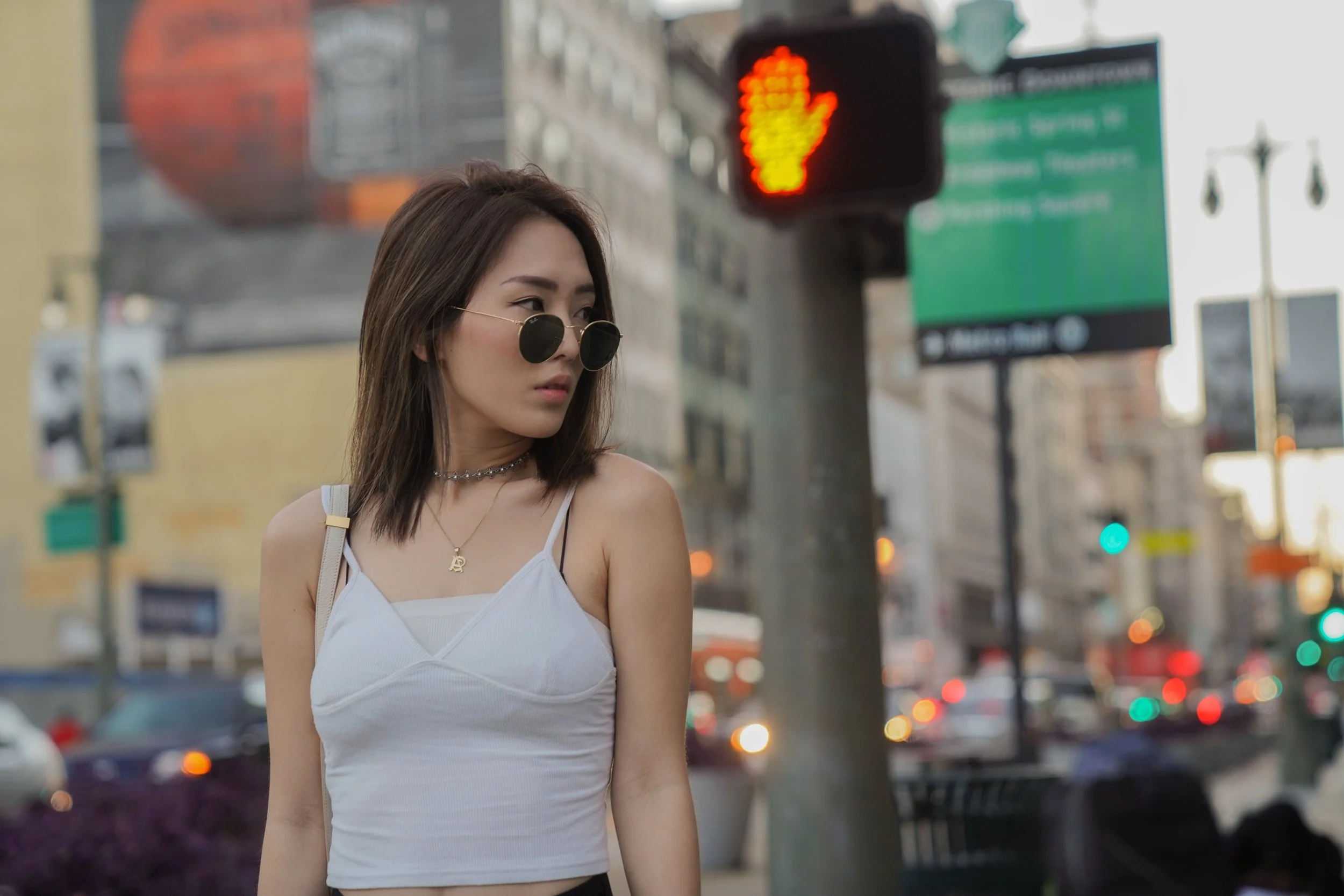 A young woman with shoulder-length brown hair wearing sunglasses, a white tank top, and layered necklaces standing on a city street with traffic, signs, and a pedestrian signal showing a red hand in the background.