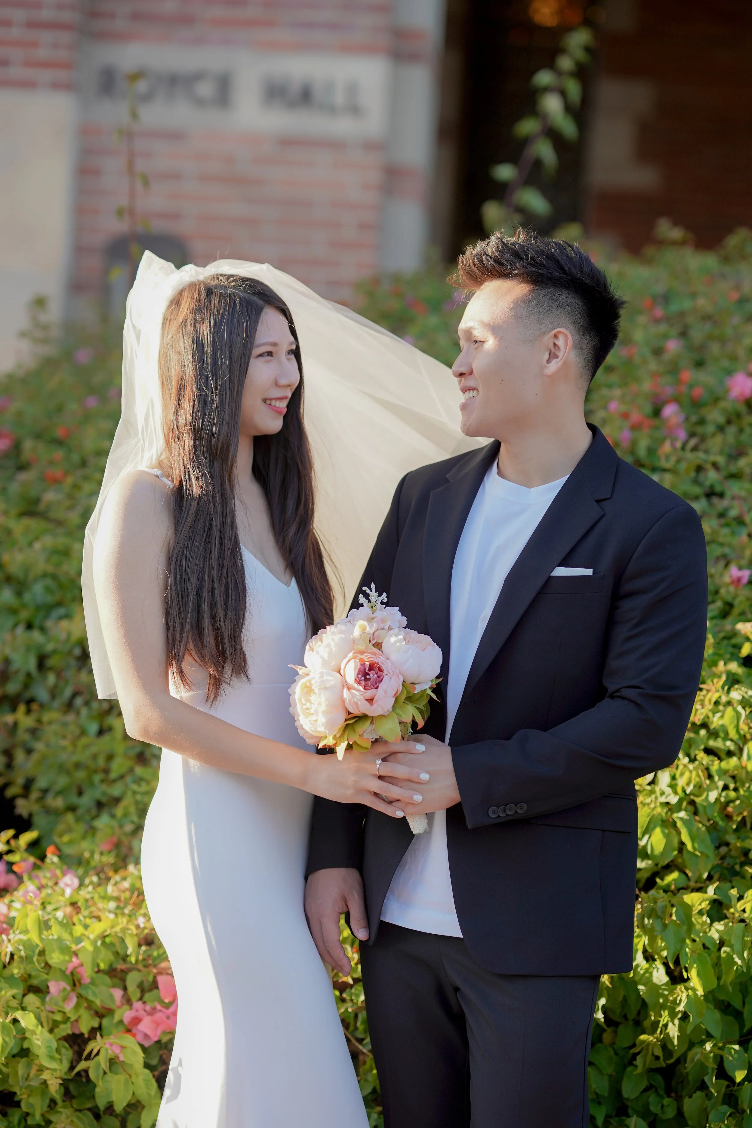 Happy bride and groom standing outdoors, holding a pink bouquet, smiling at each other.