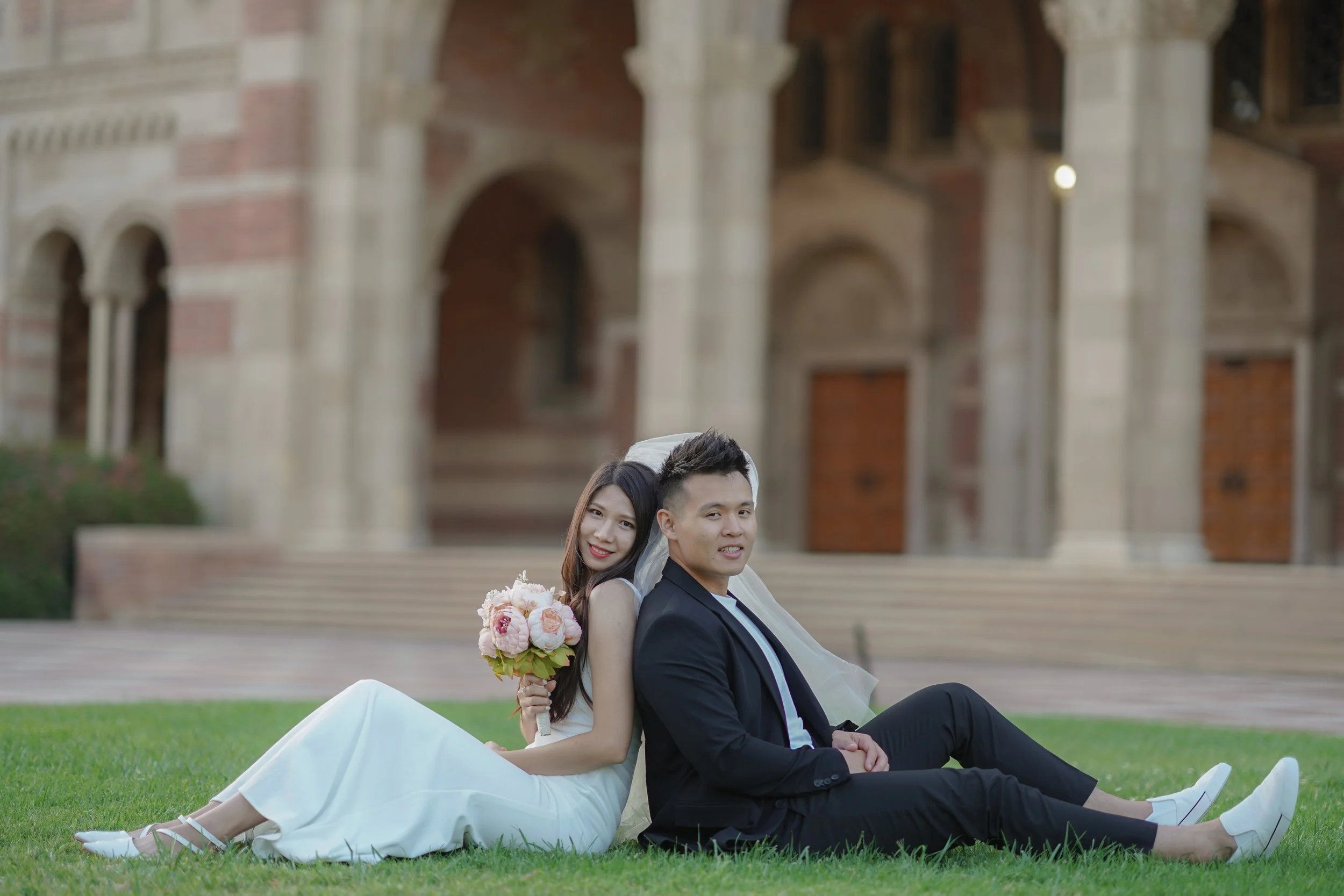 A bride and groom sit on the grass in front of a historic building, smiling and holding a bouquet of flowers.