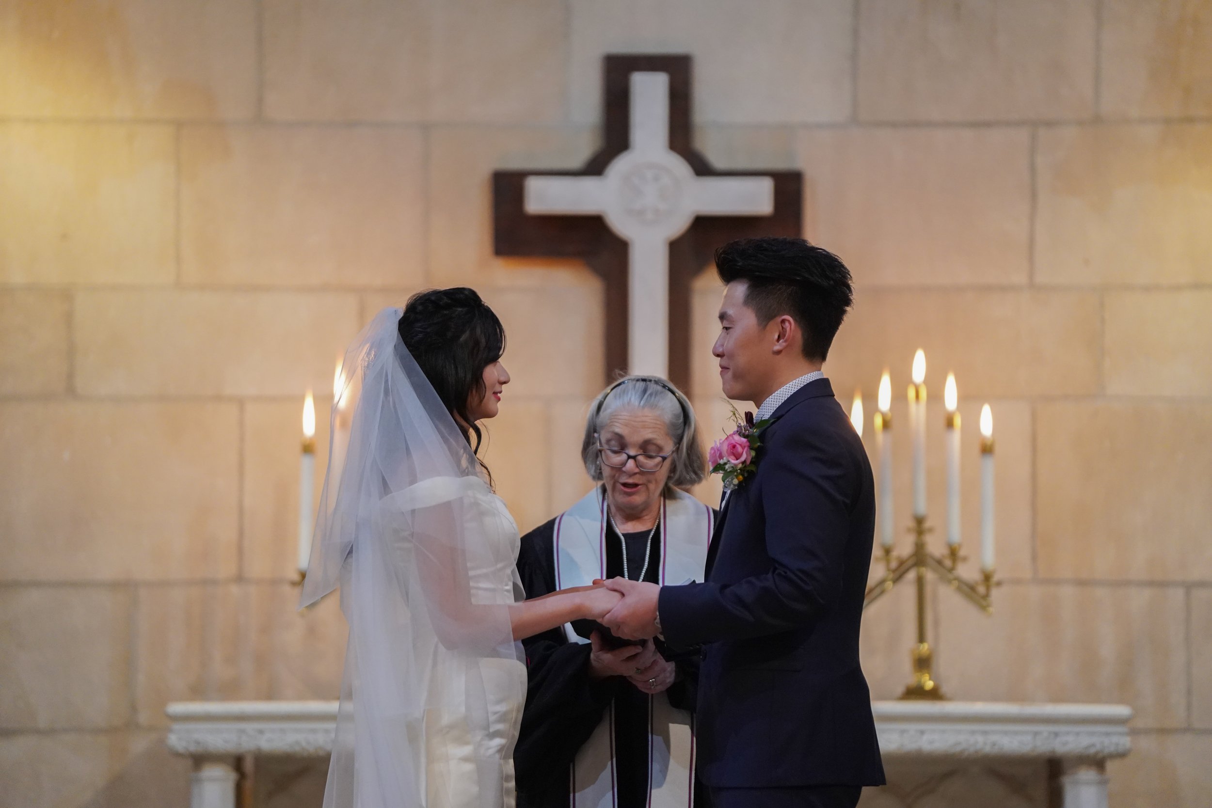 A bride and groom holding hands during their wedding ceremony in a church, with an officiant standing between them and a cross in the background.
