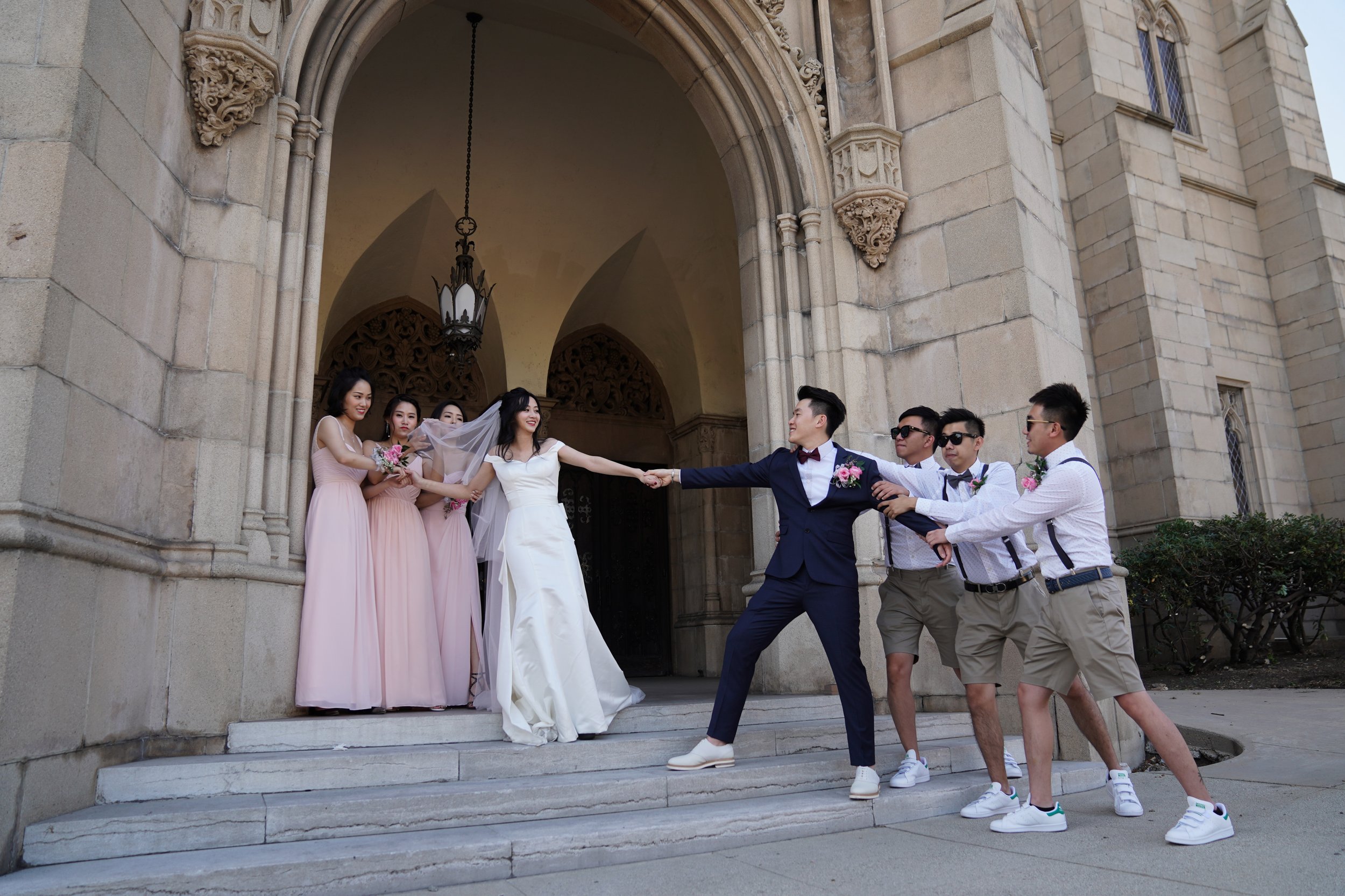 A wedding scene with the bride in a white dress and the groom in a navy suit, pulling each other's hands on the steps of a church, surrounded by bridesmaids in pink dresses and groomsmen in white shirts and shorts.