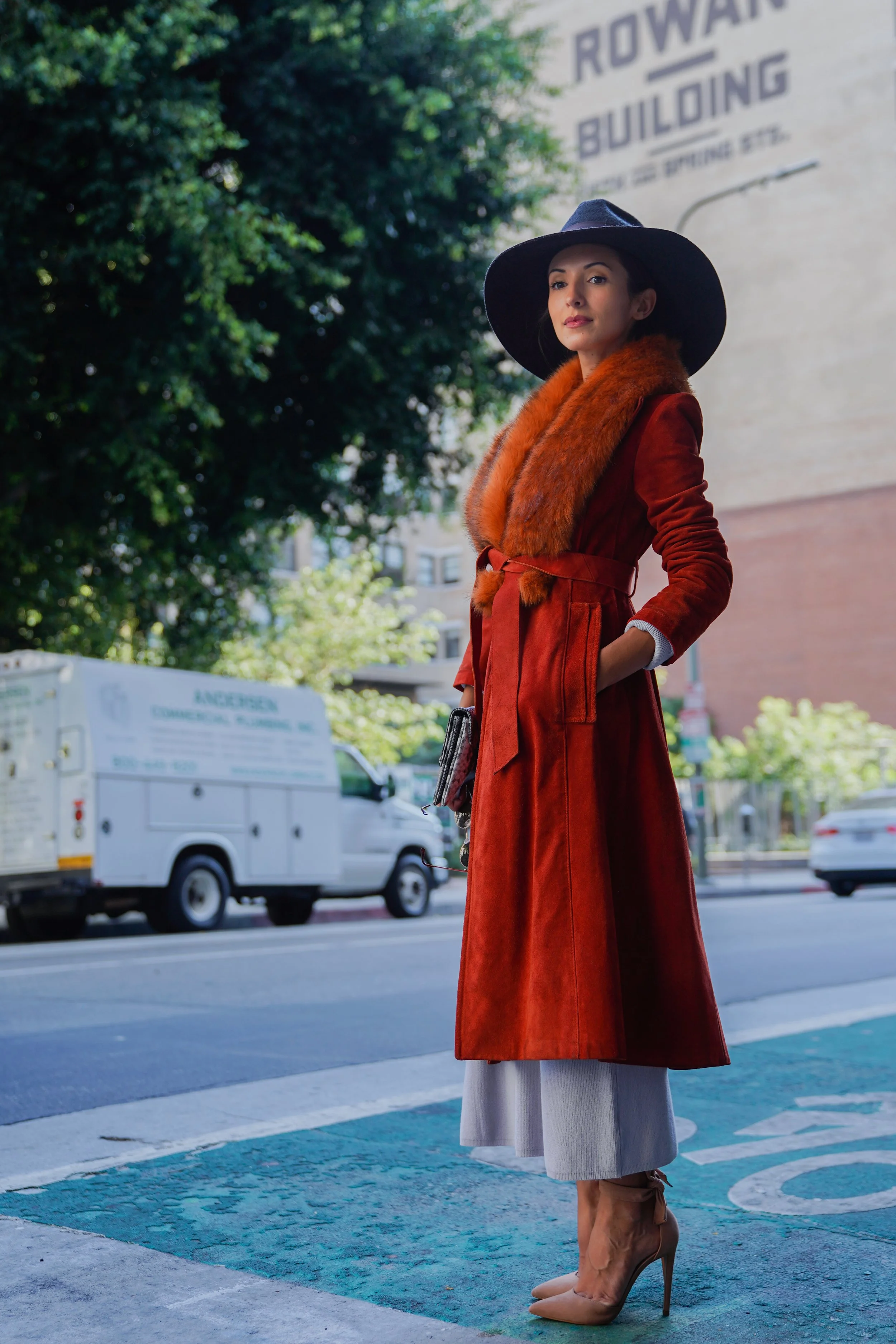 A woman standing on the sidewalk wearing a red coat, a large black hat, tan high heels, and a fur scarf, with trees and cars in the background.