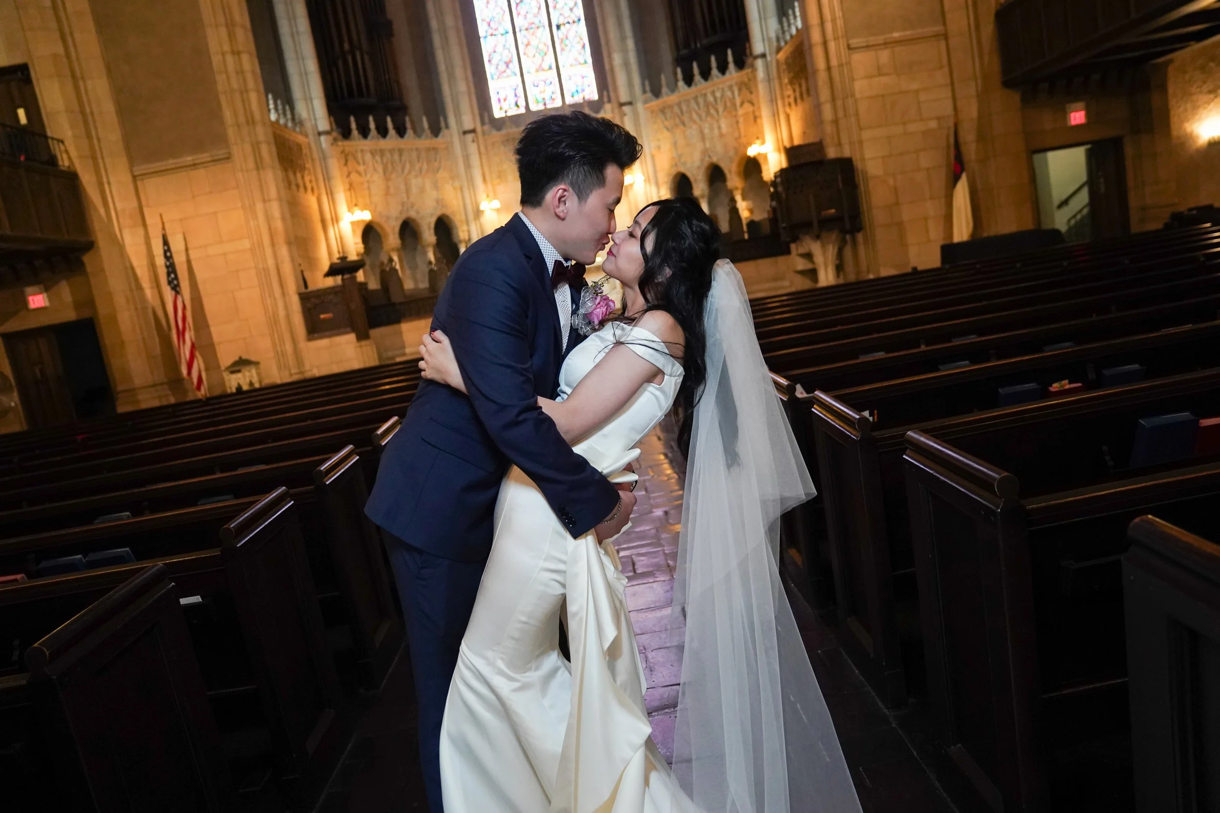 A bride and groom share a kiss inside a church with wooden pews and stained glass windows.