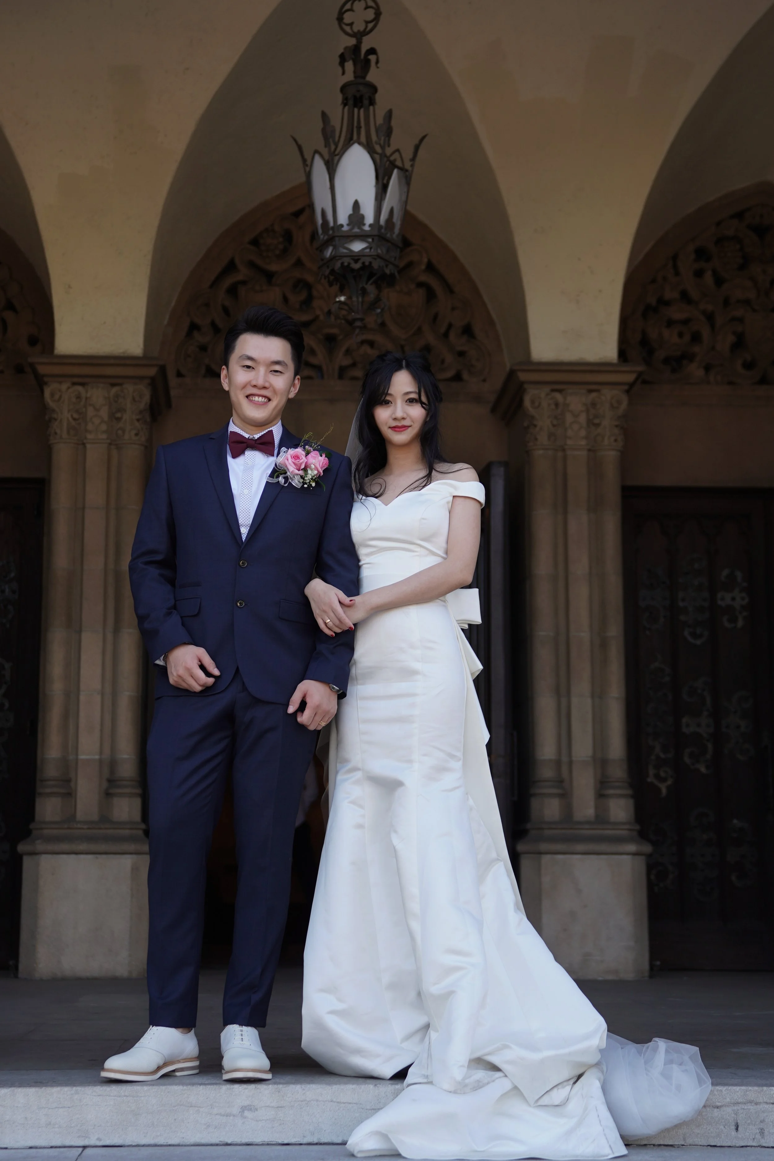 A bride and groom standing outside a building with intricate arches, smiling for a wedding photo. The groom is wearing a navy blue suit with a pink boutonniere, and the bride is in a white wedding gown with a bow at the back.