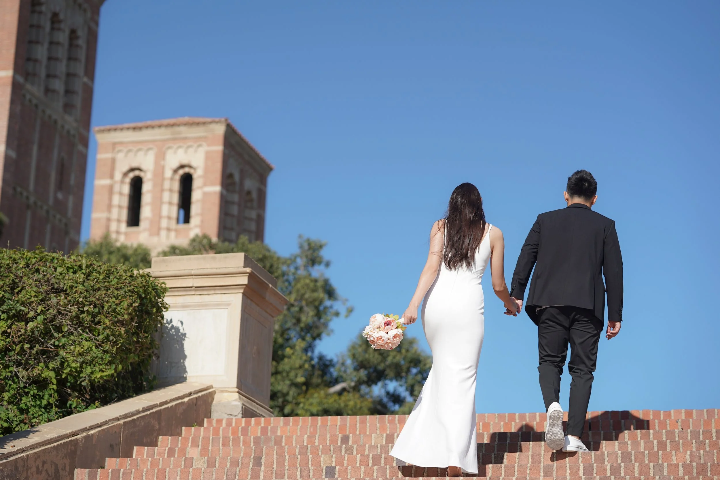 A bride in a white wedding dress holding a bouquet walking hand-in-hand with a groom in a black suit up brick stairs outdoors on a sunny day.
