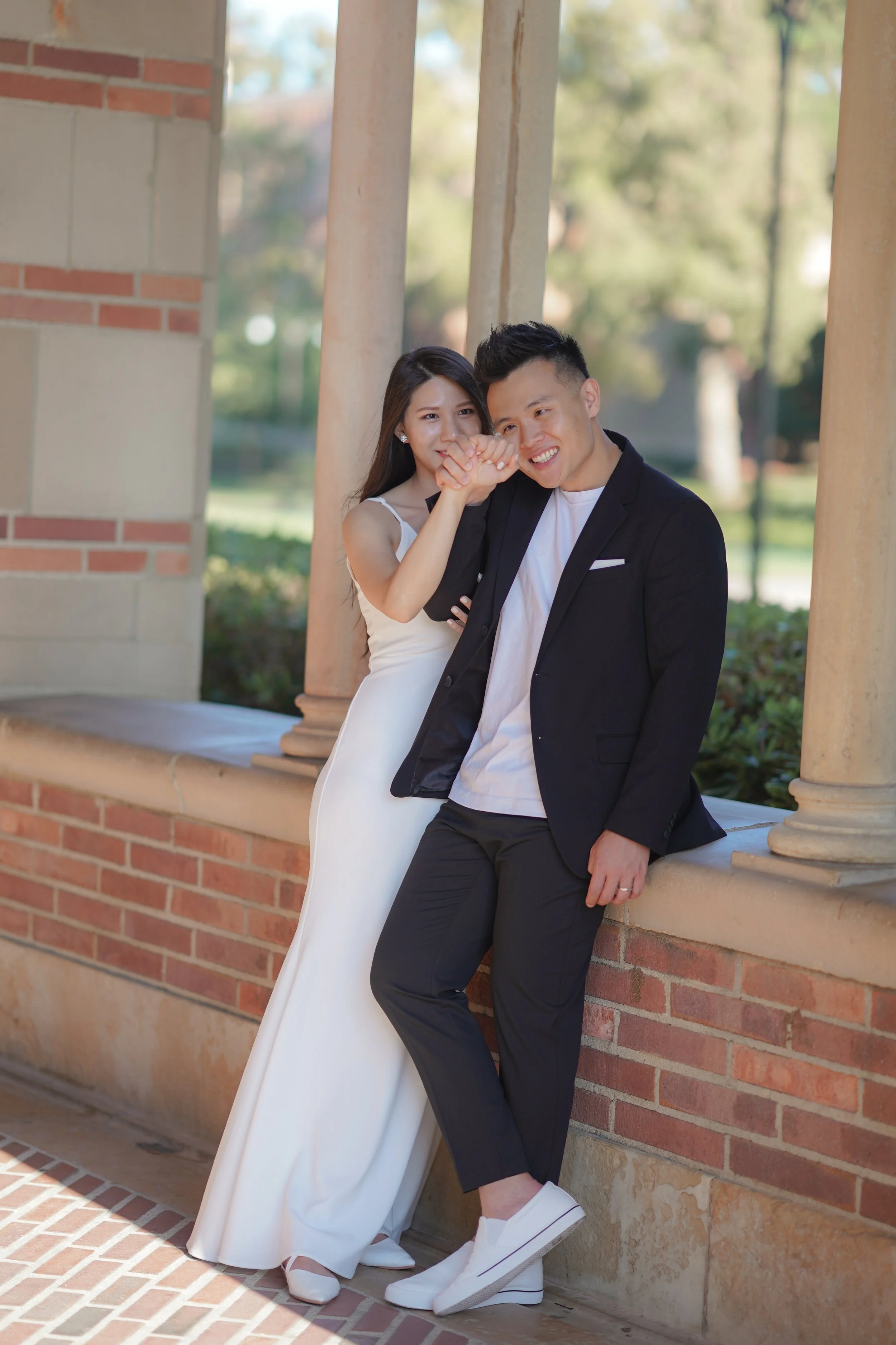 A smiling couple, dressed in formal attire, leaning on a brick wall with columns outdoors on a sunny day.