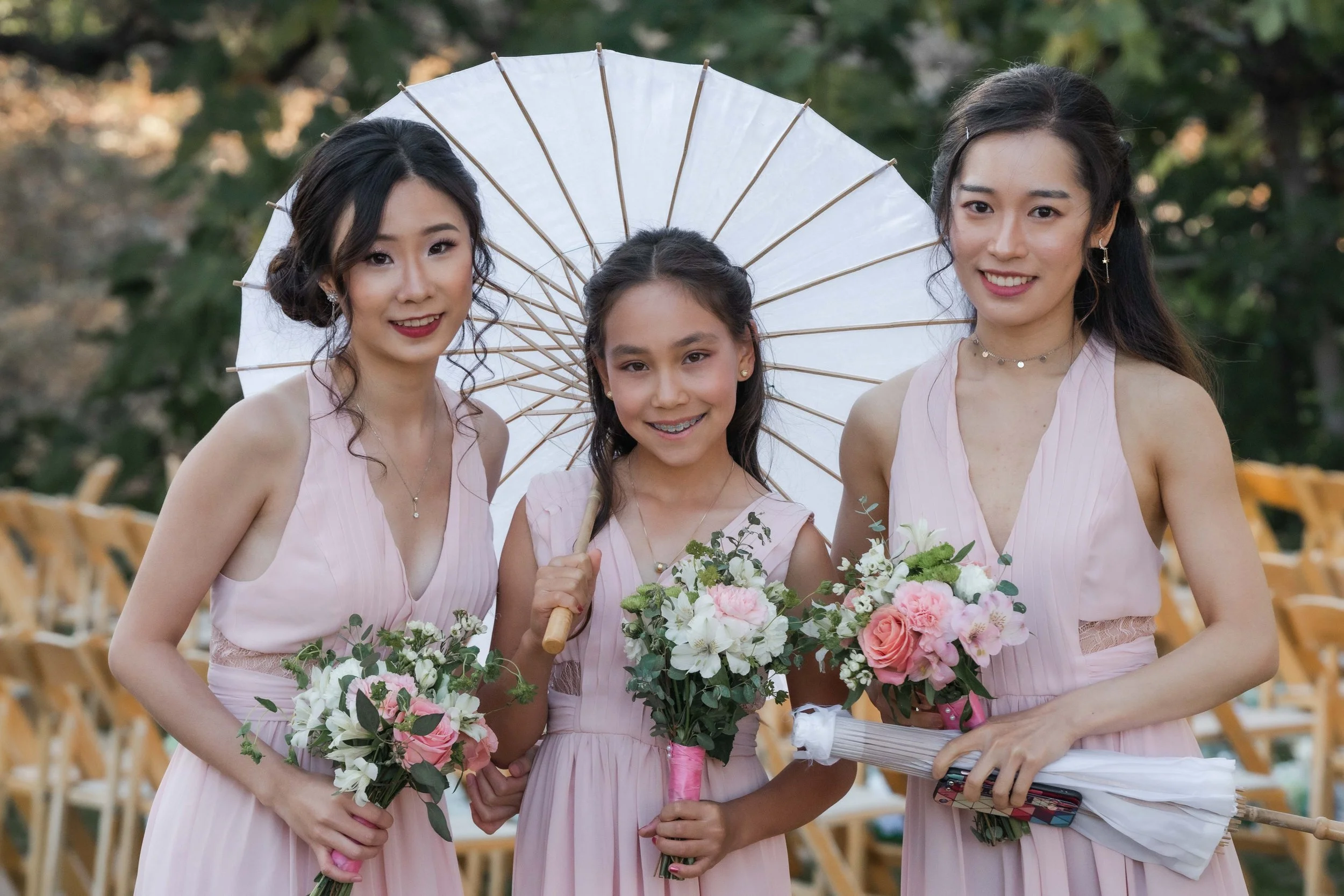 Three young women in pink dresses at an outdoor event, holding bouquets of flowers. One woman is holding a white parasol. They are smiling and standing in front of a background of chairs and greenery.