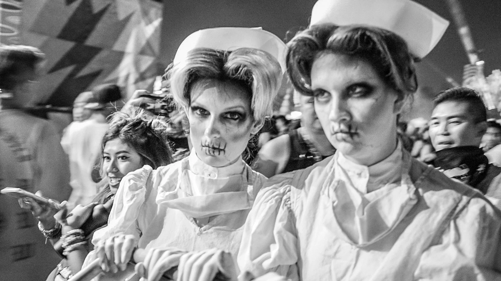 Two people dressed in skeleton makeup and costumes, with short, styled hair, attending a nighttime event with a crowd around them.