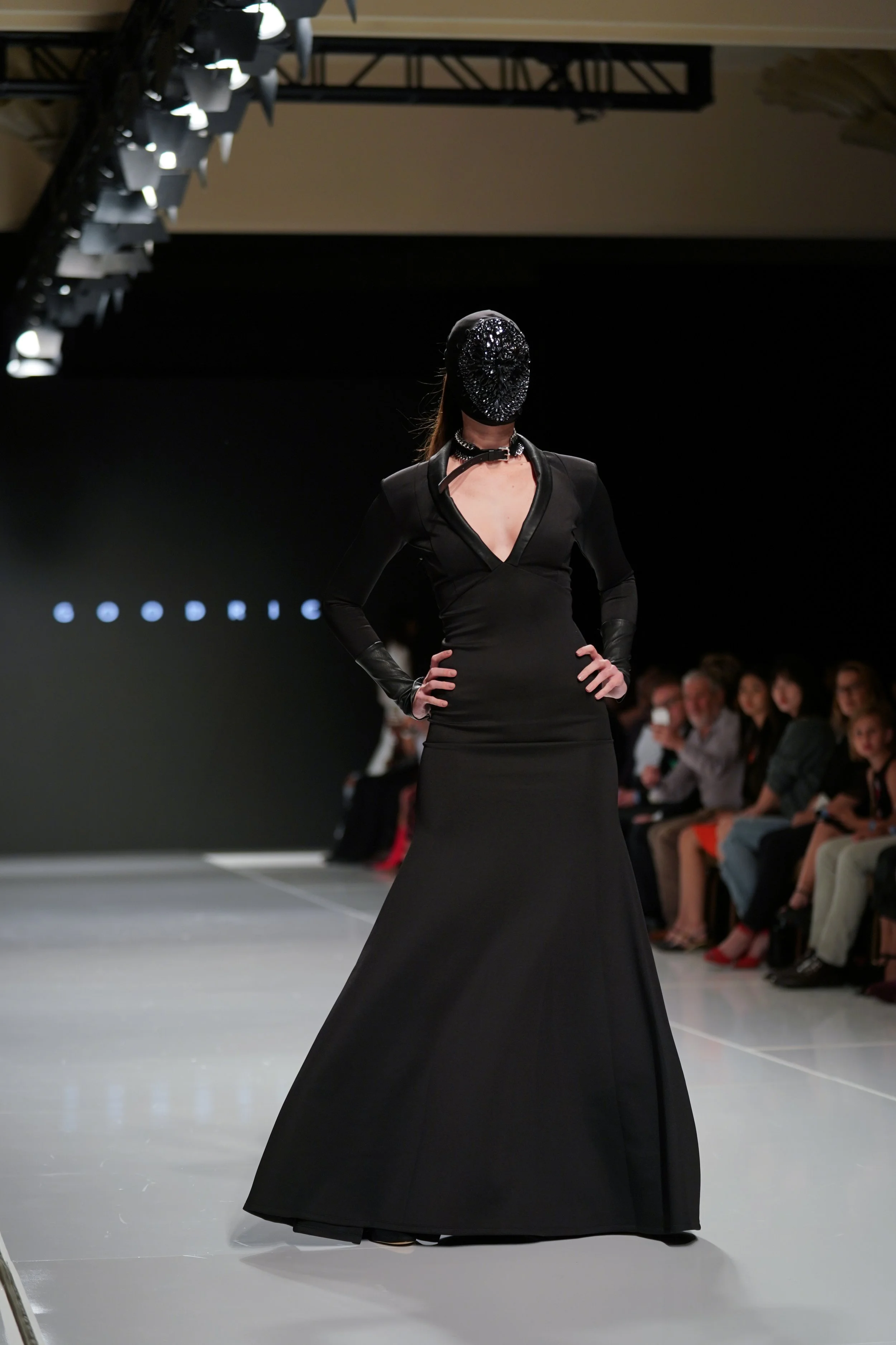 Model walking down a runway during a fashion show, wearing a black long-sleeved gown with a deep V-neck and a decorative black headpiece. Audience members are seated along the runway watching the show.