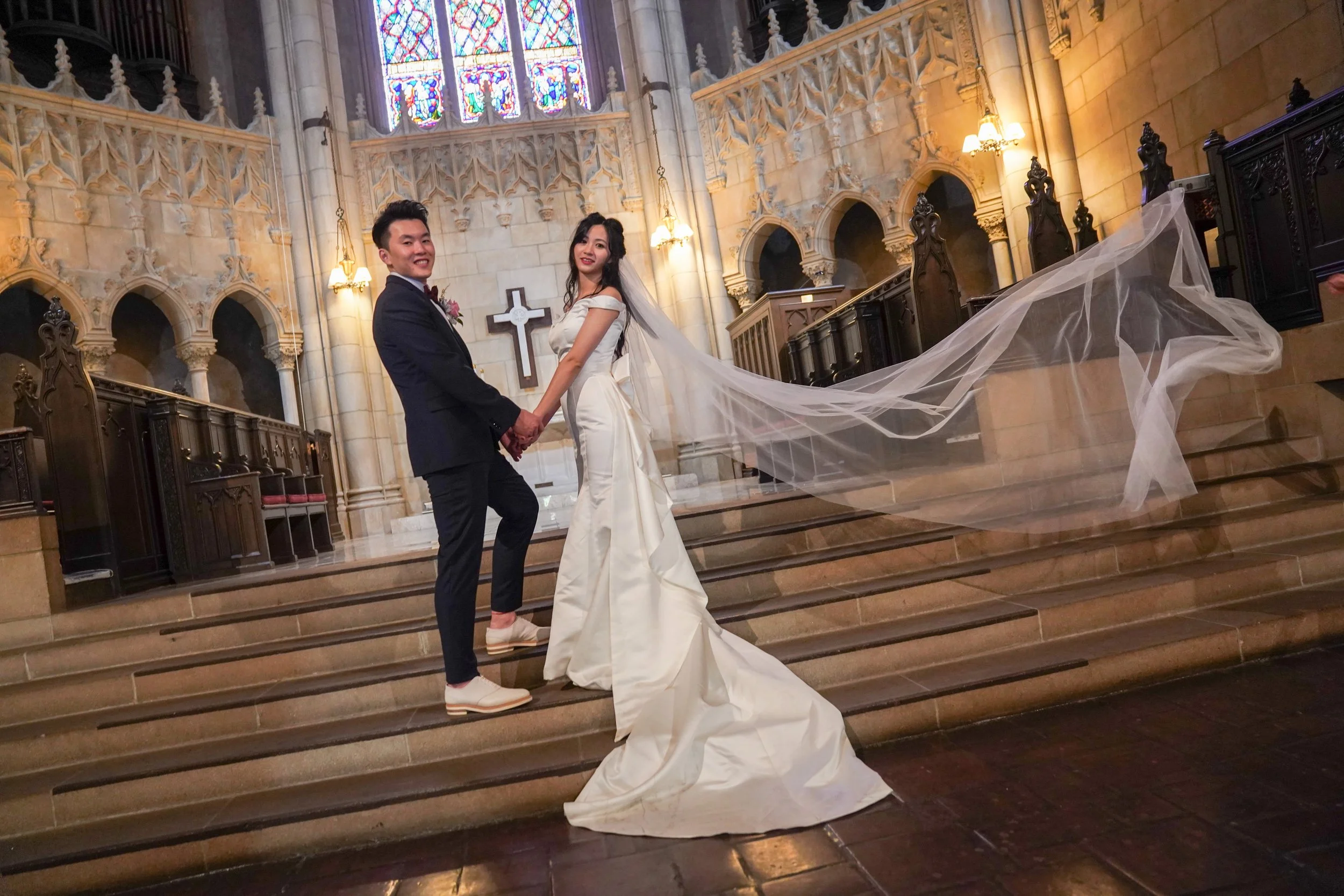 A bride and groom holding hands on church stairs, with stained glass windows and a cross in the background. The bride is wearing a long white dress with a veil, and the groom is in a dark suit with white shoes.
