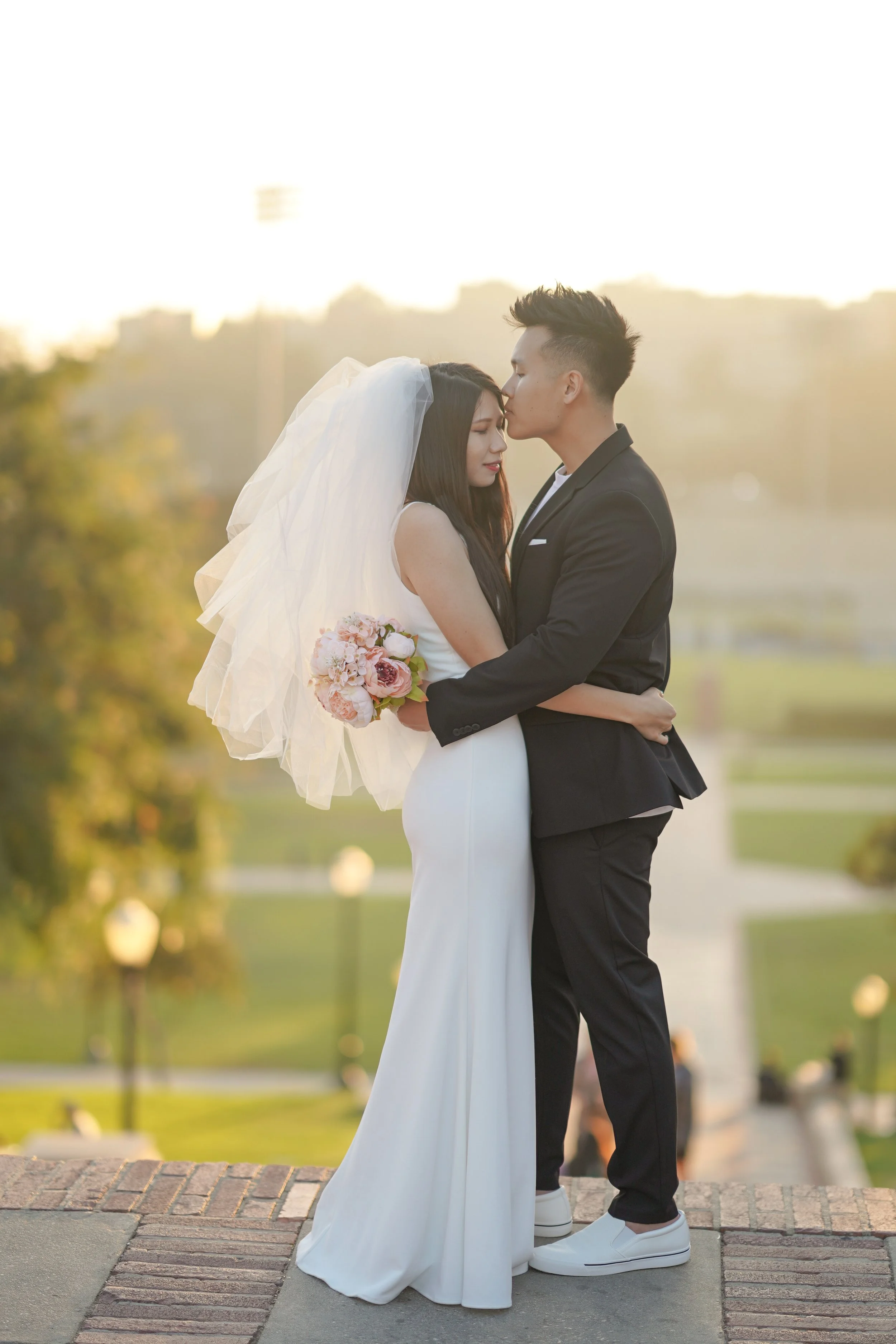 A bride and groom embrace outdoors at sunset, with the groom giving a gentle kiss on the bride's forehead. The bride wears a white gown and veil, holding a pink and white bouquet, while the groom is dressed in a black suit and white sneakers.