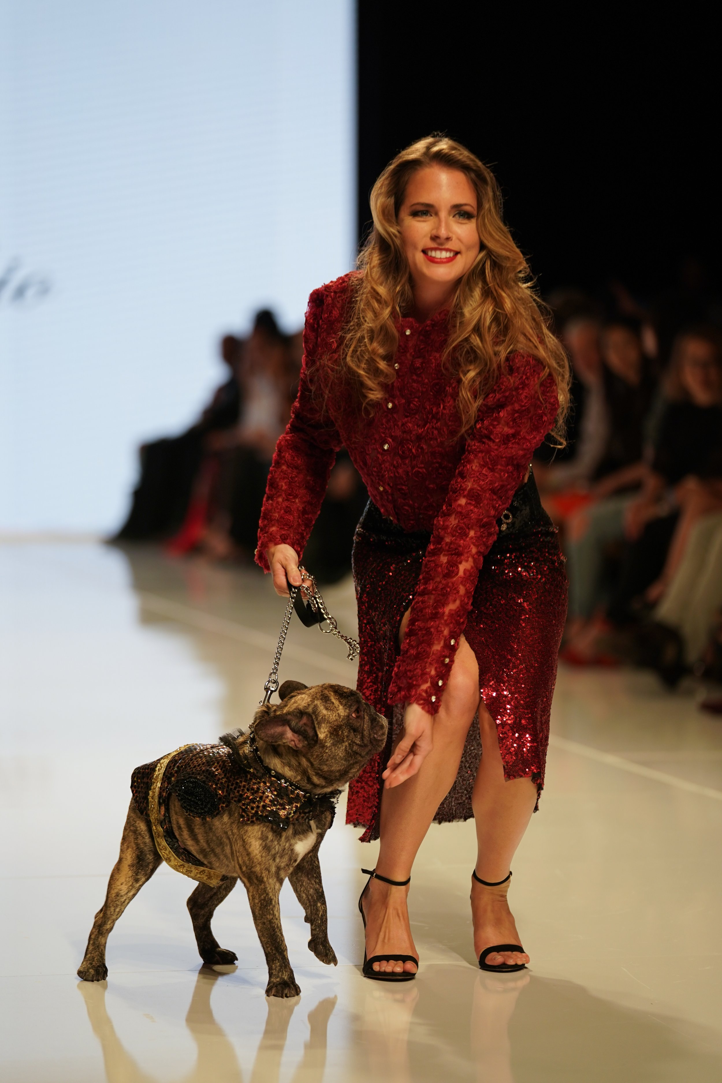 A woman with long curly hair in a red, sequined dress walking a brindle dog on a runway during a fashion show.