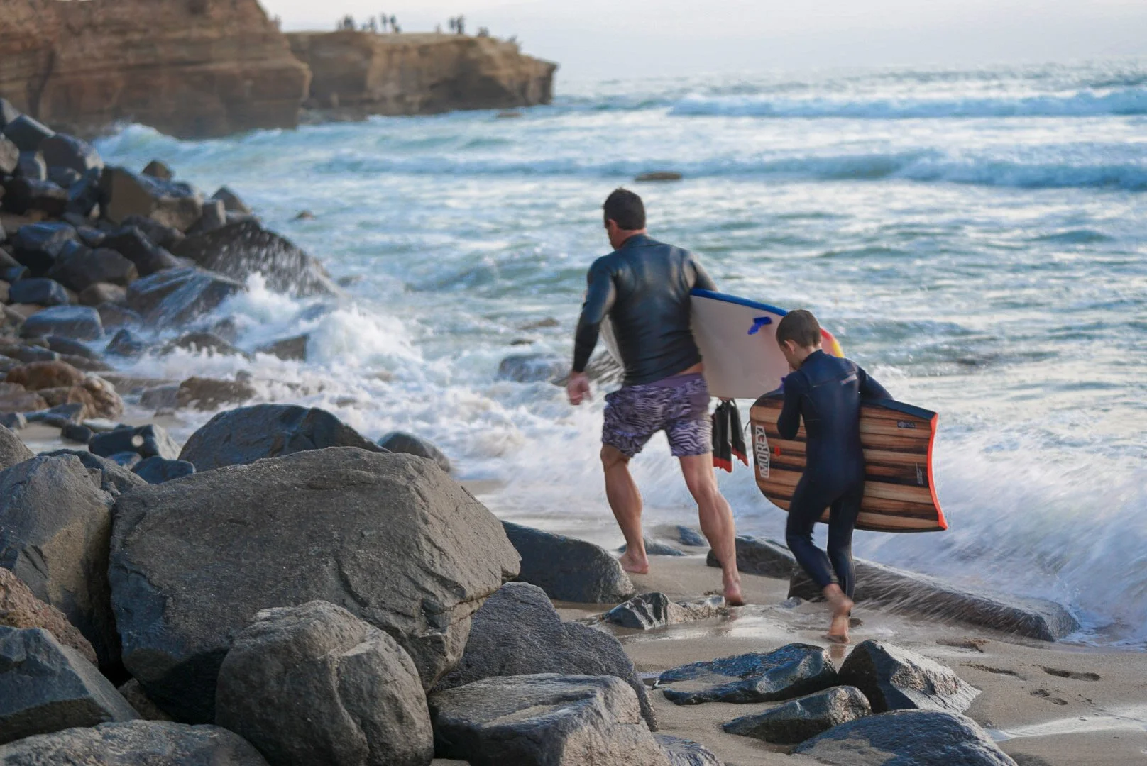 A man and a boy carrying surfboards walk onto the beach through rocks, heading towards the ocean.
