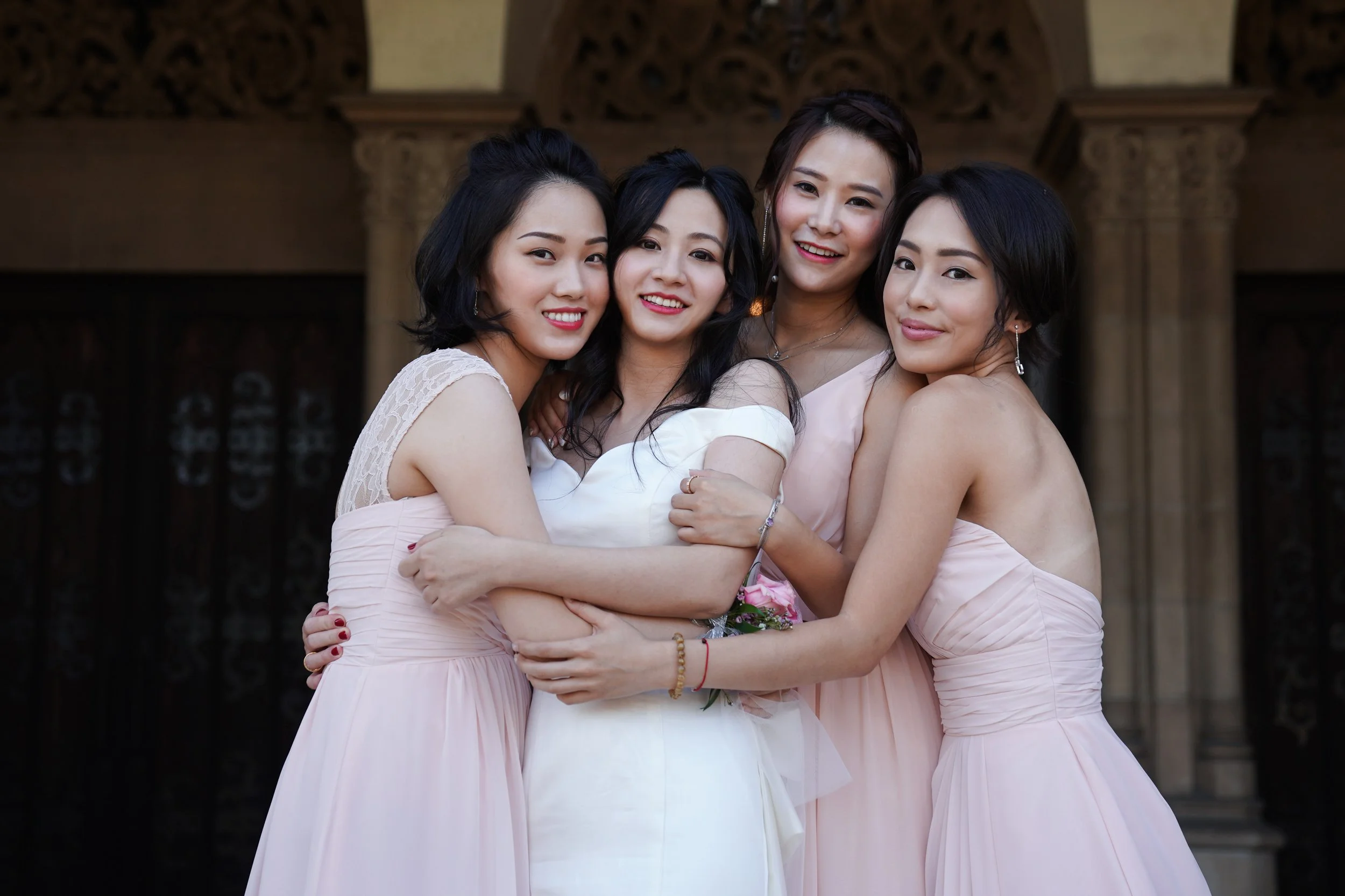 Four women dressed in pastel-colored formal gowns embracing each other and smiling for the camera, standing outdoors in front of a decorative architectural background.