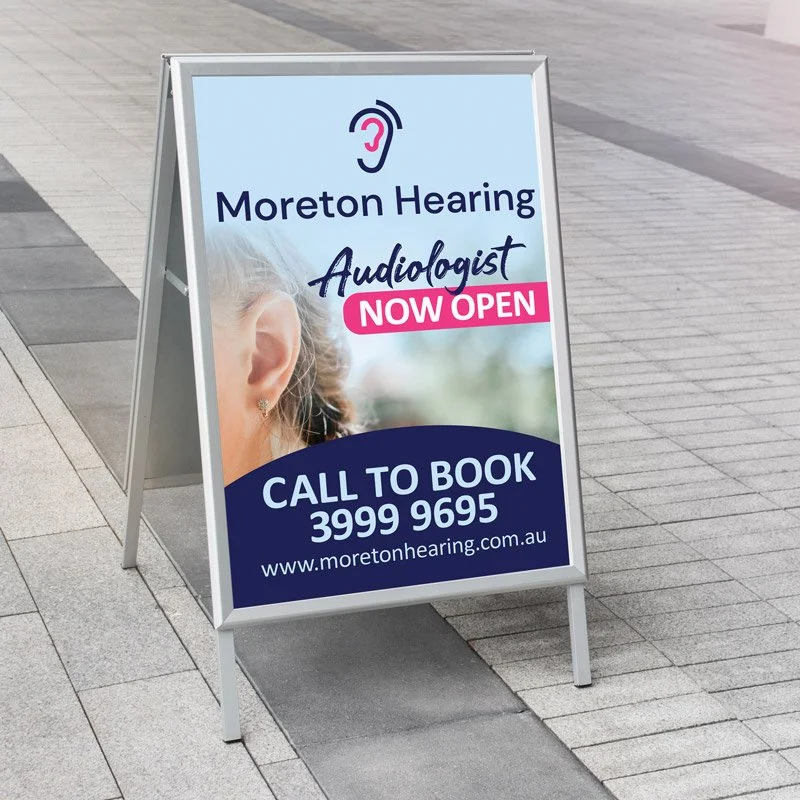 A-frame footpath sign for Moreton Hearing featuring a close-up image of an ear. The sign includes the text “Moreton Hearing Audiologist – Now Open” in navy and pink lettering