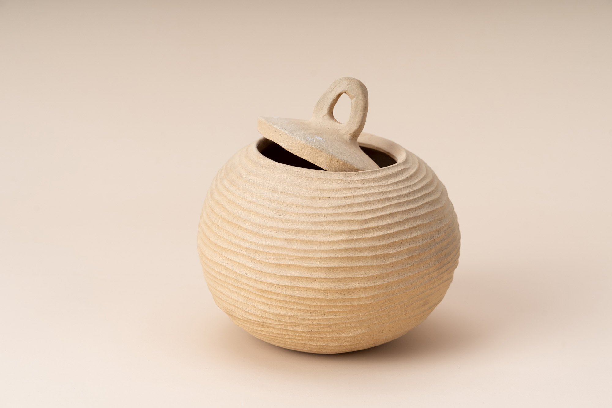 A beige, round ceramic jar with a ribbed texture and a lid that is partially open, showing the interior.