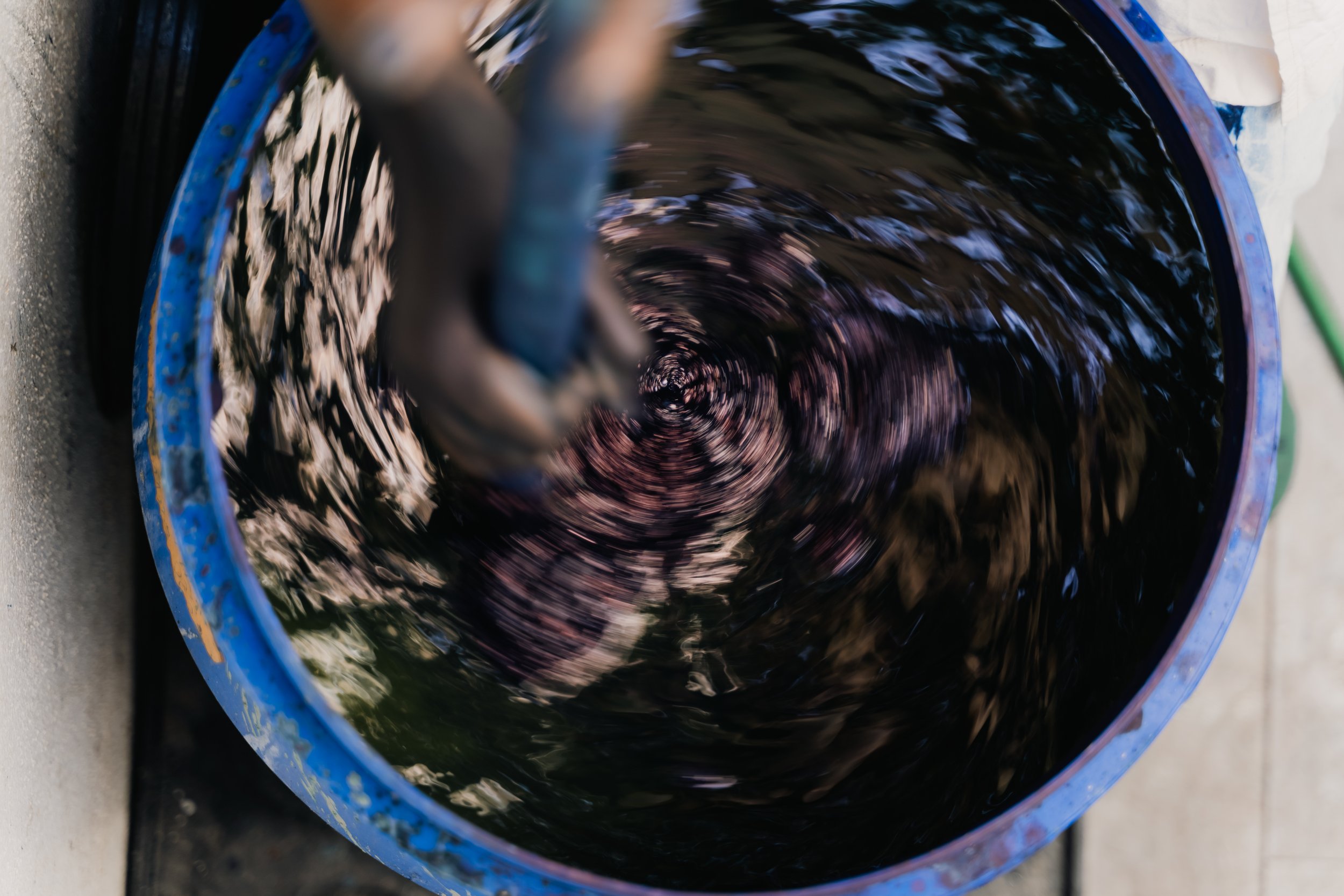 A person stirring a dark liquid in a blue bucket with a paintbrush.